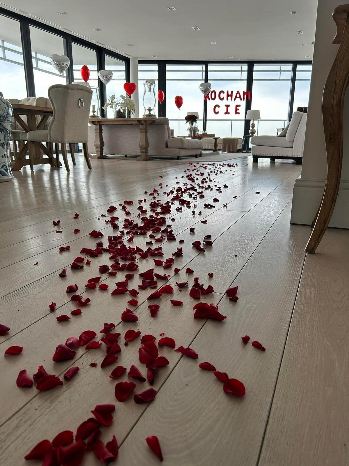 Romantyczna aranżacja wnętrza: płatki czerwonych róż rozsypane na jasnej podłodze prowadzące do salonu z balonami w kształcie serc i napisem 'KOCHAM CIE'.