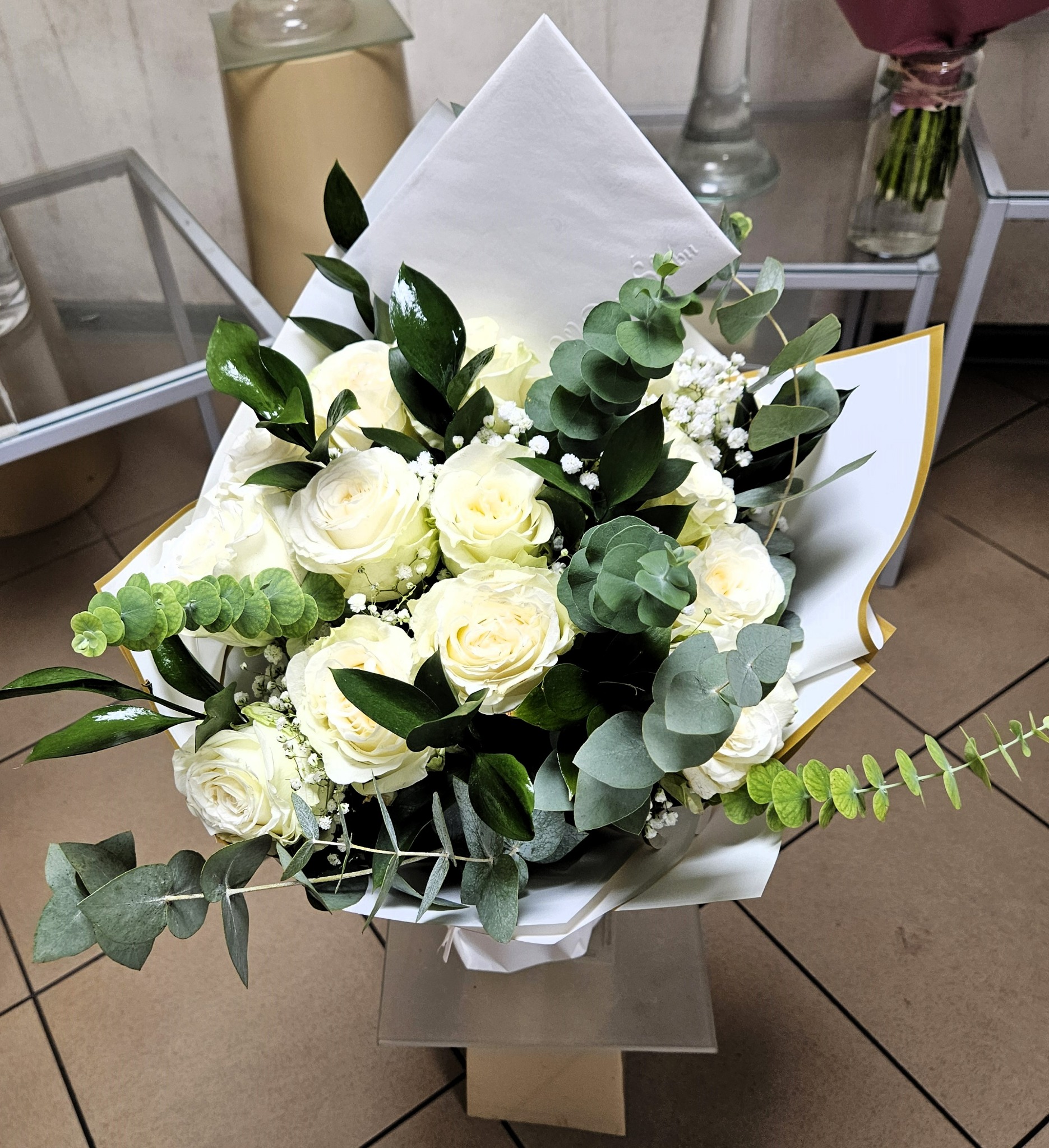 Elegancki bukiet z białych róż, eukaliptusa i gipsówki, zapakowany w biały papier ze złotym obramowaniem, umieszczony na szklanym stoliku.