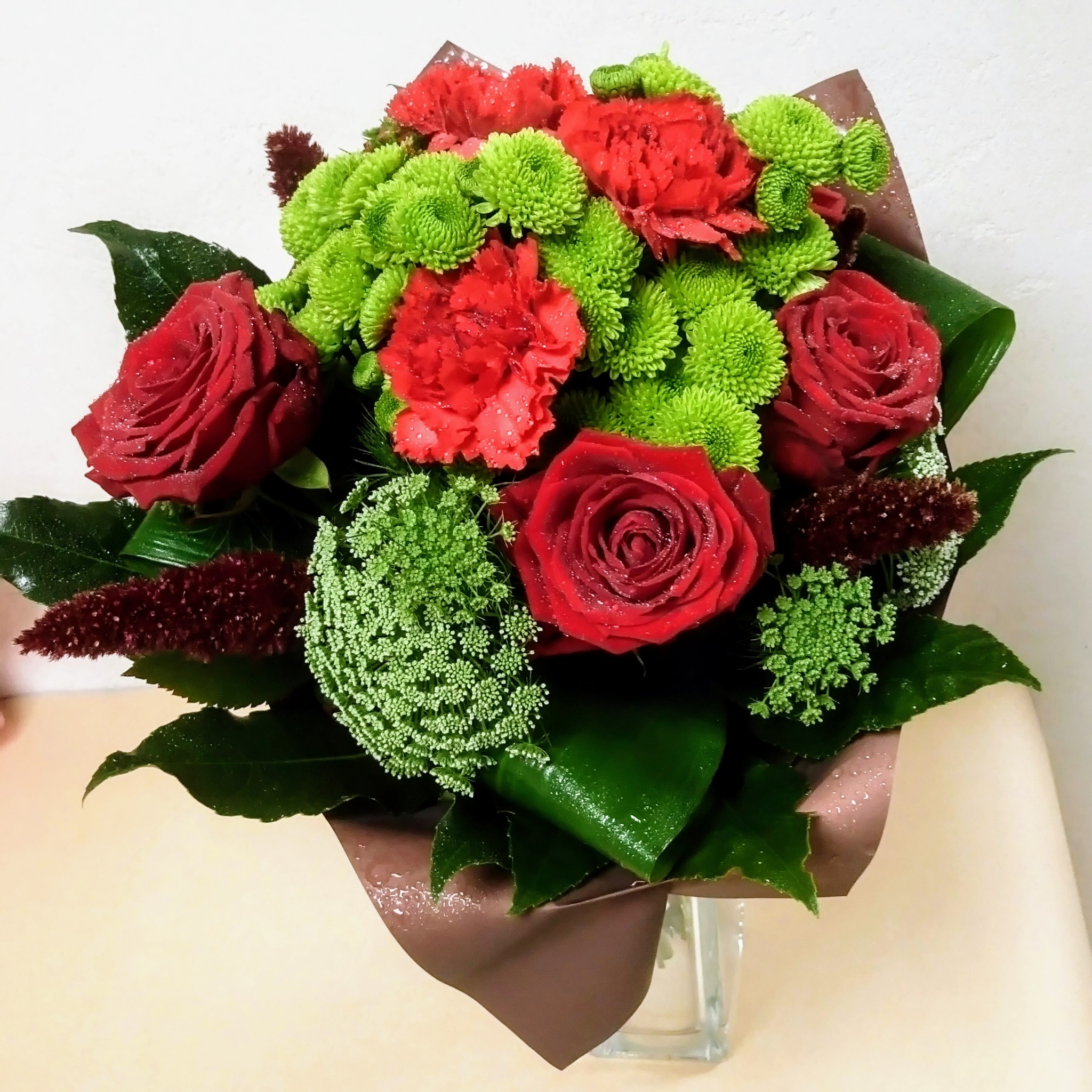 Bukiet czerwonych róż, goździków i zielonych chryzantem w brązowym papierze, z kroplami wody na płatkach, umieszczony w szklanym wazonie.