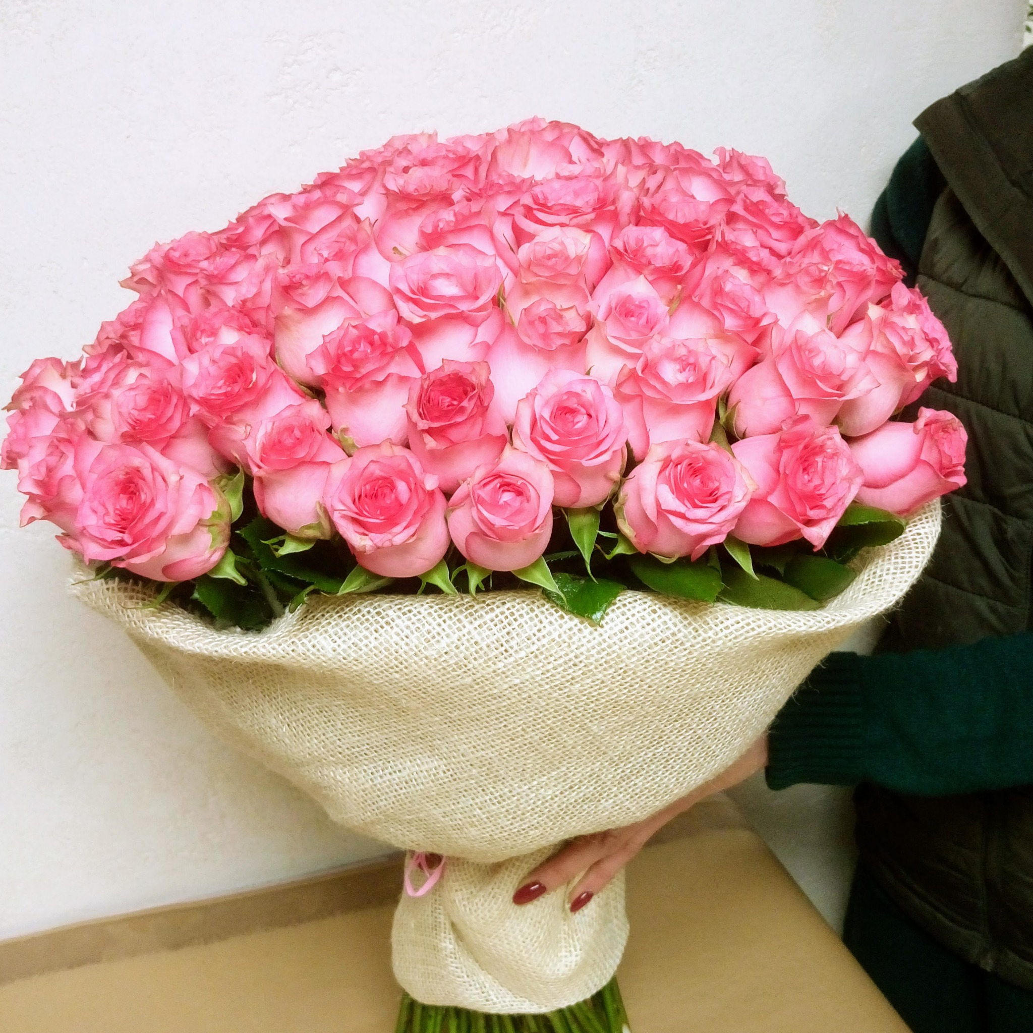 Duży bukiet różowych róż z białymi brzegami, owinięty w jutowy materiał i przewiązany różową wstążką, trzymany przez osobę w zielonym swetrze i kamizelce.