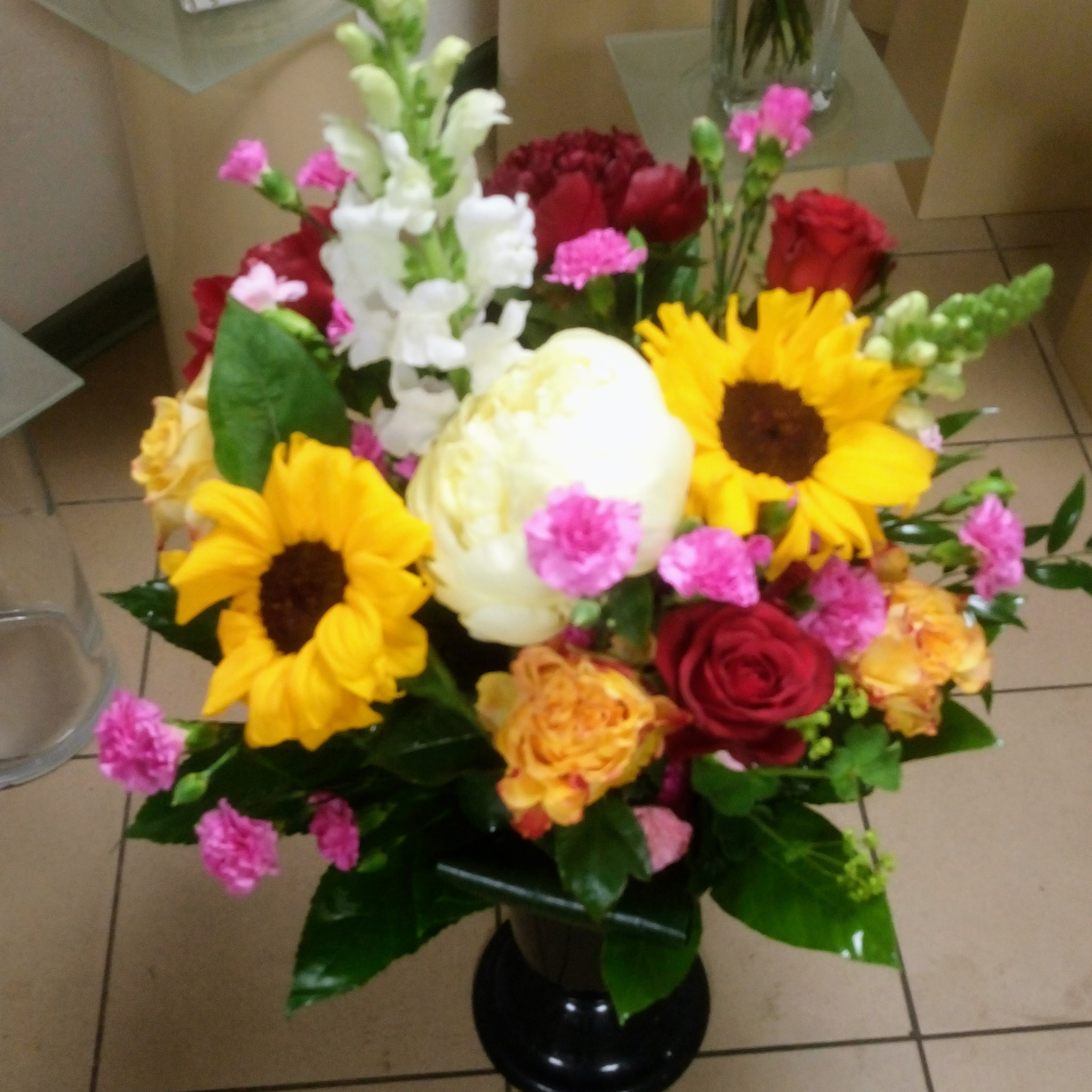 Kompozycja kwiatowa z żółtych słoneczników, białych piwonii, czerwonych i pomarańczowych róż oraz różowych goździków w czarnym, okrągłym wazonie na tle beżowych płytek podłogowych.