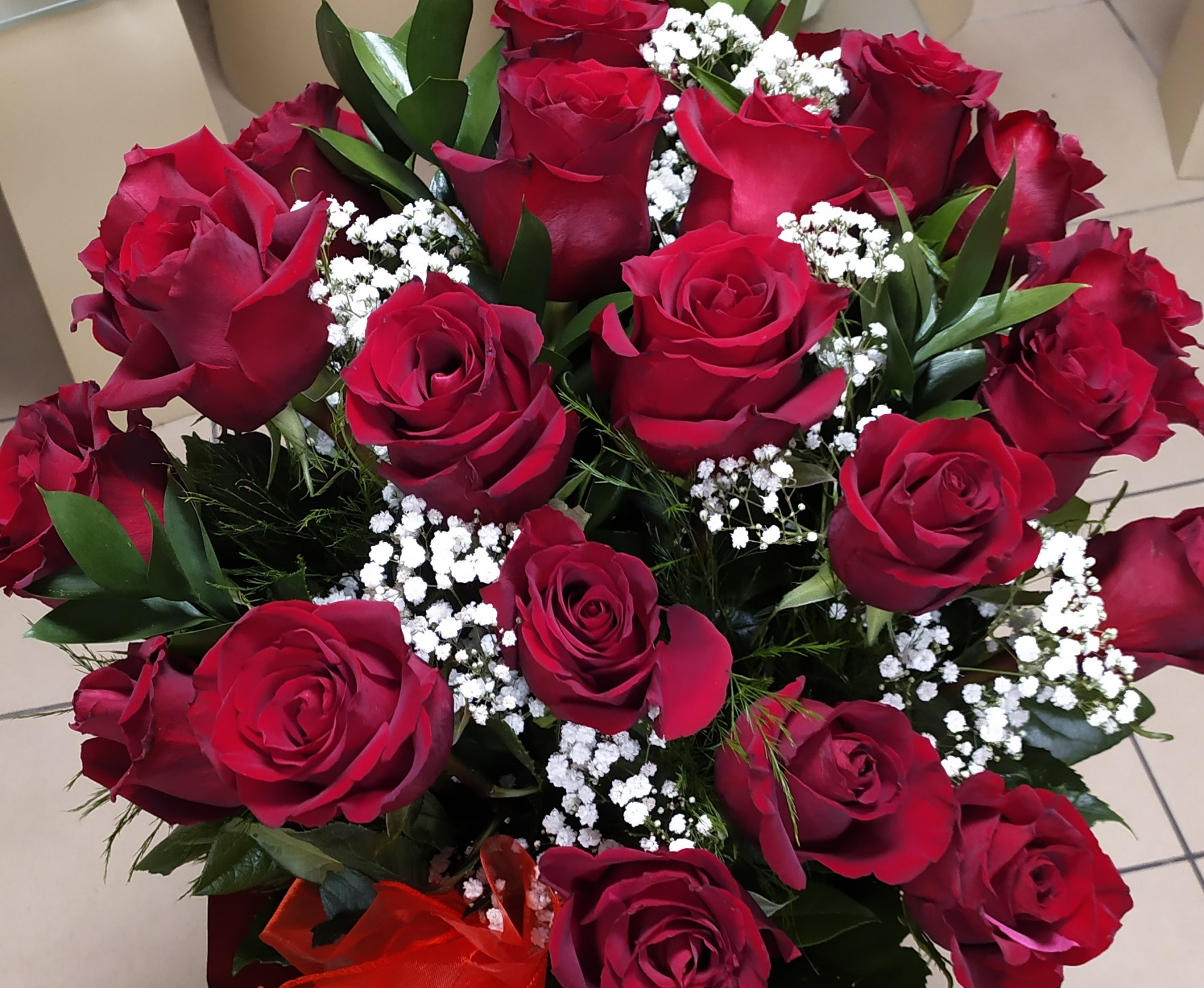 Bukiet ciemnoczerwonych róż z białymi gipsówkami i zielonymi liśćmi, przewiązany czerwoną wstążką, na tle jasnej podłogi z płytek.