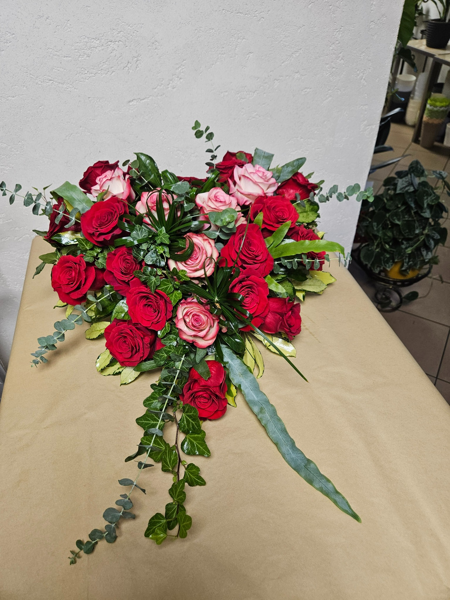 Kompozycja kwiatowa z czerwonych i różowych róż, eukaliptusa i bluszczu na beżowym tle, widoczna doniczka z zieloną rośliną w tle.