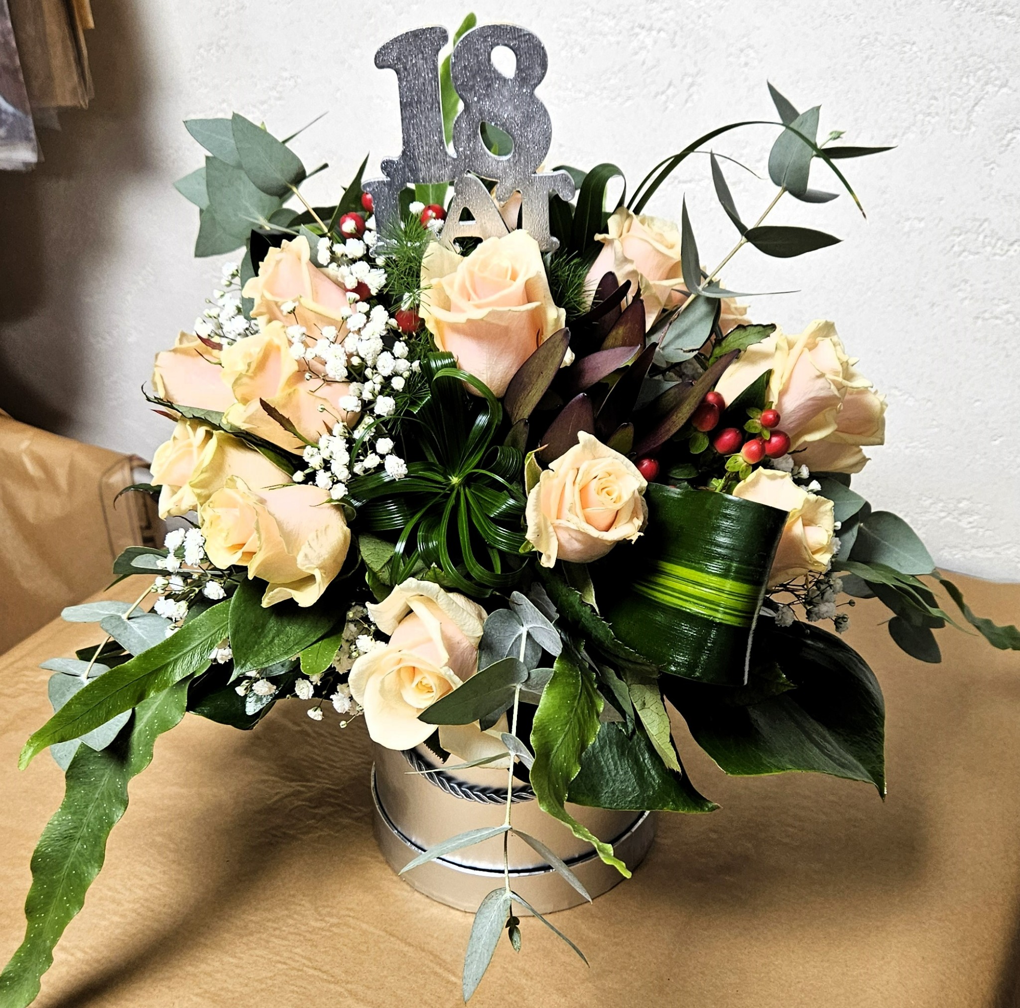 Kremowe róże w srebrnym pudełku z dekoracją '18 lat', uzupełnione zielonymi liśćmi eukaliptusa, drobnymi białymi kwiatkami i czerwonymi jagodami.