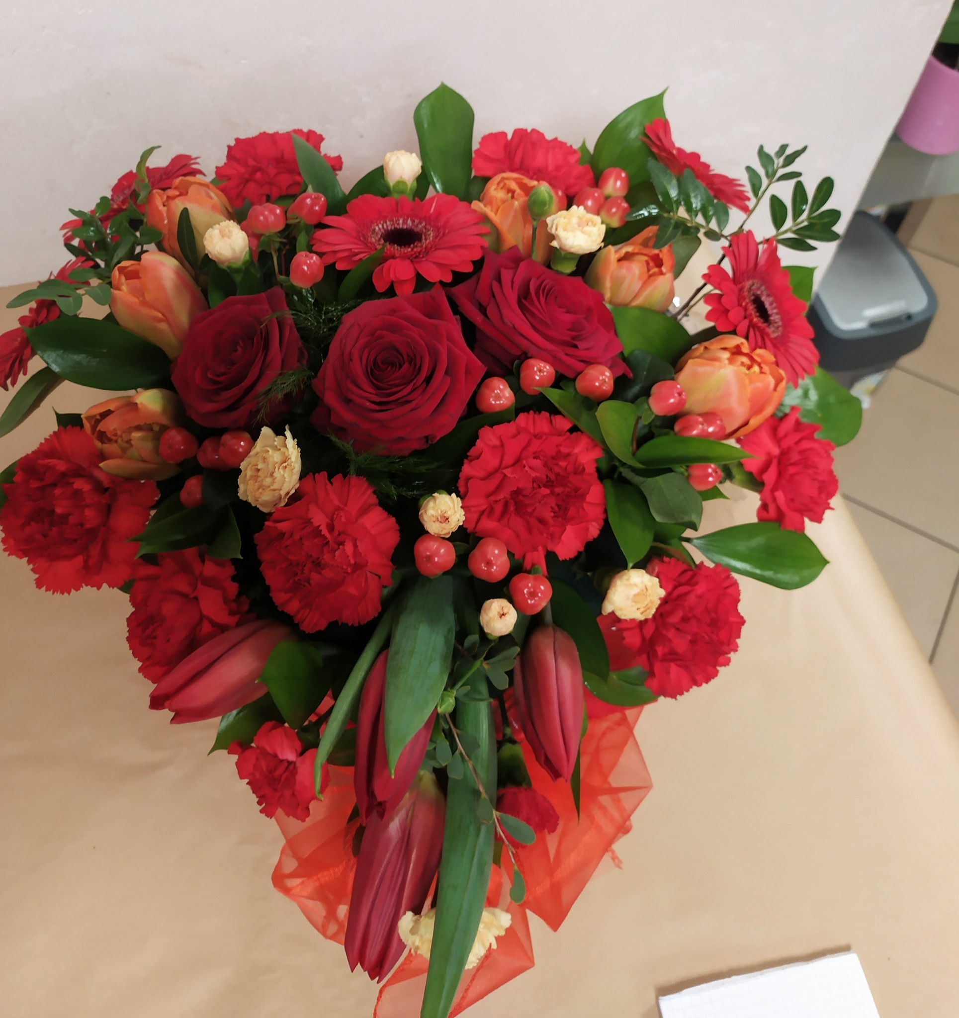 Kompozycja kwiatowa w odcieniach czerwieni i pomarańczy, z różami, goździkami, gerberami i tulipanami, udekorowana czerwonym tiulem, na jasnym tle.