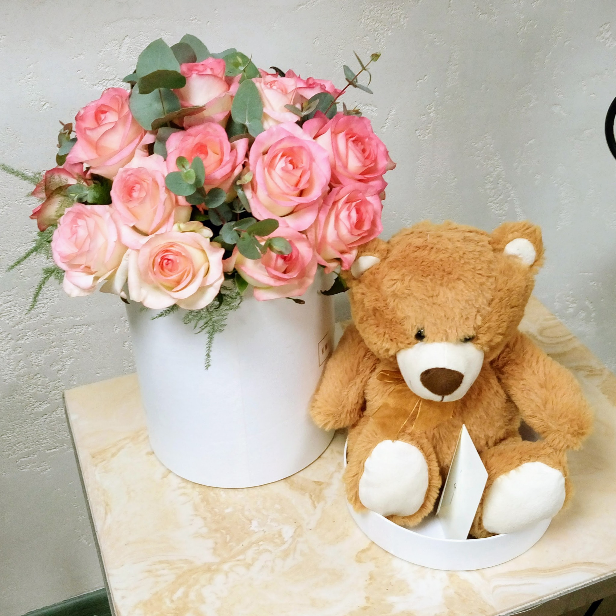 Kompozycja z pudrowych róż i eukaliptusa w białym pudełku, obok siedzi pluszowy miś z białą kartką.