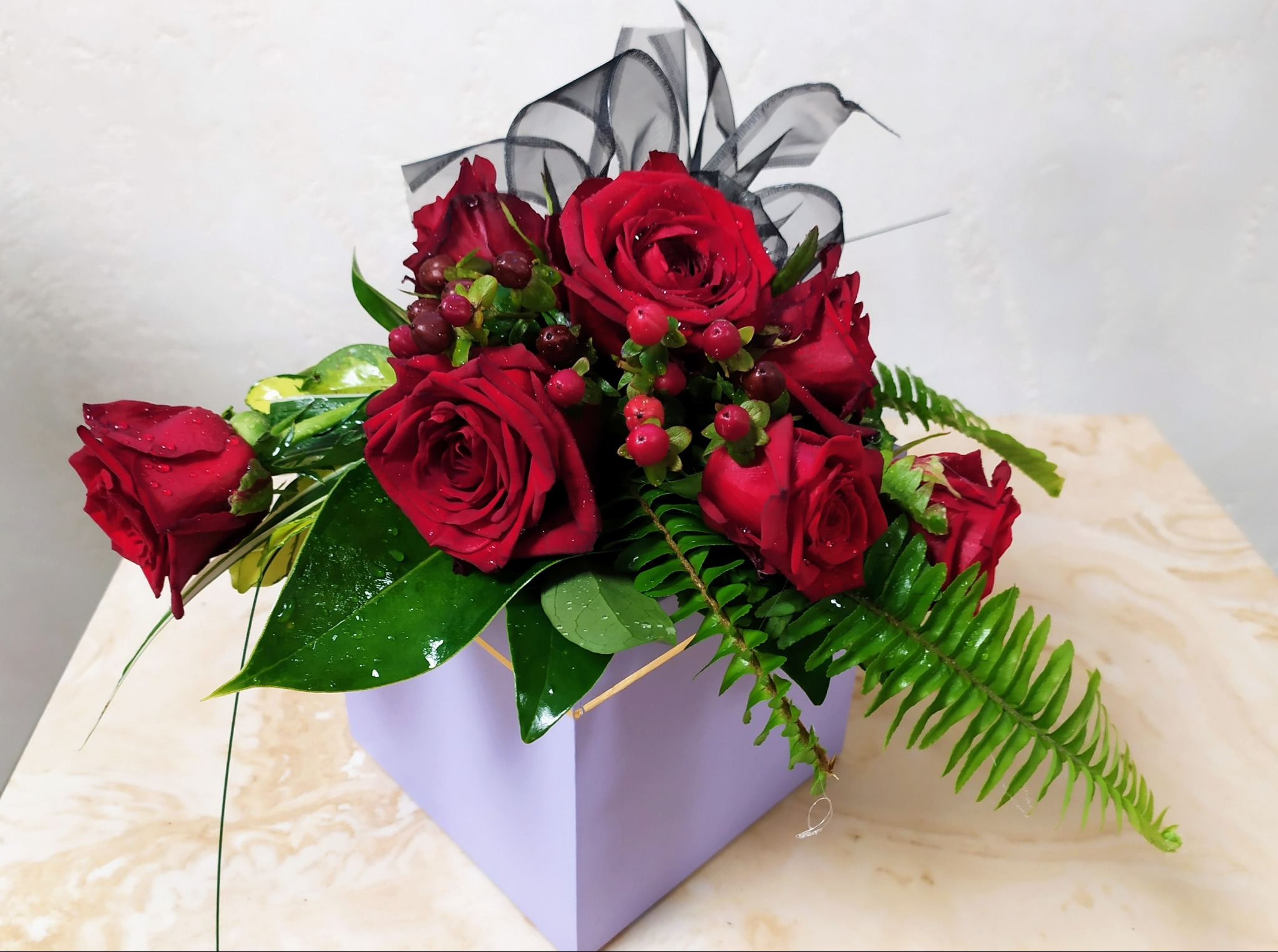 Kompozycja z czerwonych róż, zielonych liści i czarnej wstążki w liliowym, kwadratowym pudełku na marmurowym blacie.