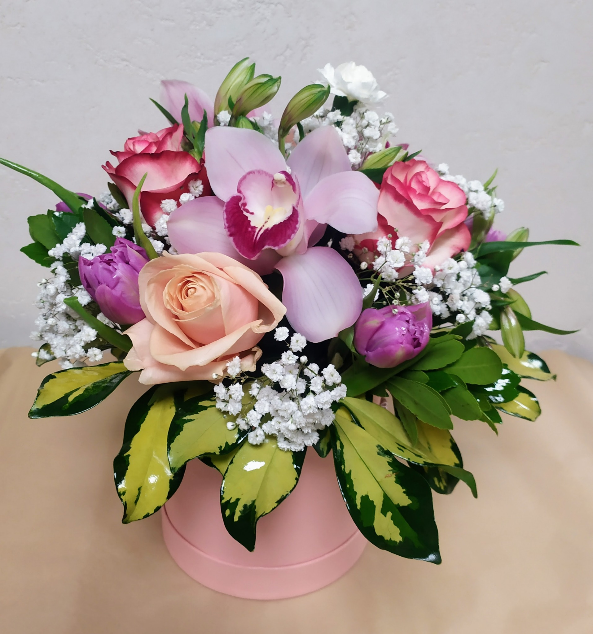 Kompozycja kwiatowa w pudelku: różowe róże z obwódką, liliowe tulipany, jasnoróżowa orchidea i gipsówka, z liśćmi o żółto-zielonych brzegach.