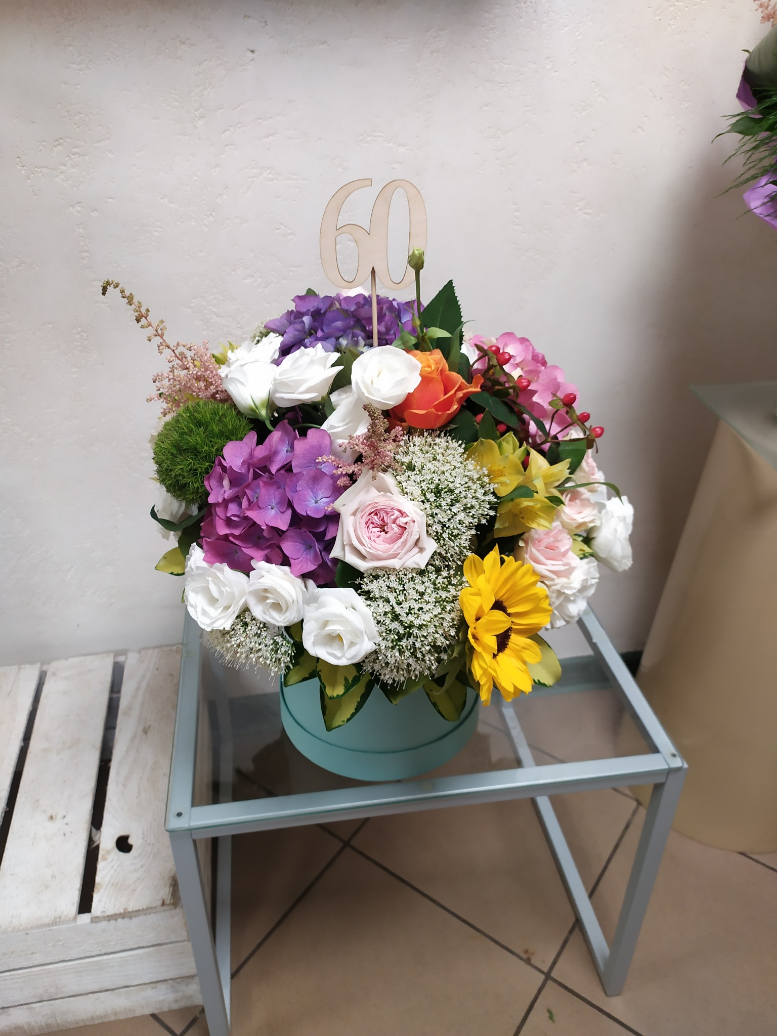 Kompozycja kwiatowa w błękitnym pudełku z drewnianym numerem 60, zawierająca hortensje, róże, słonecznik i drobne białe kwiaty, umieszczona na szklanym stoliku.