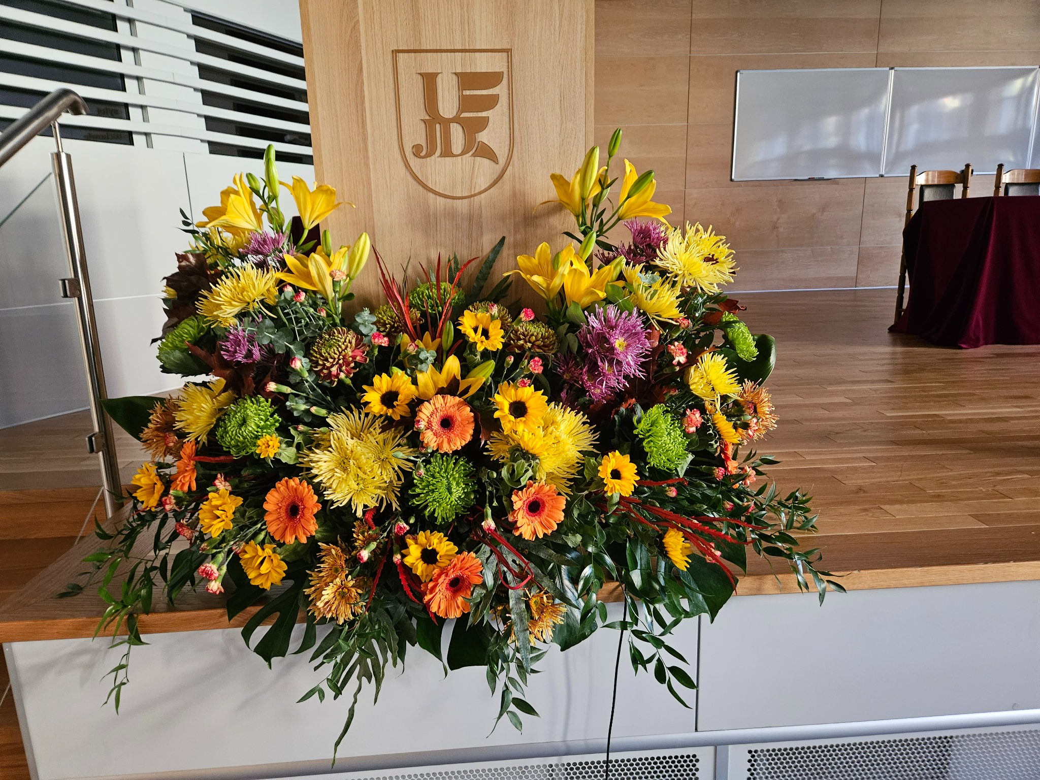 Dekoracja kwiatowa z lilii, gerber i chryzantem w odcieniach żółci, pomarańczy i fioletu na drewnianym podeście z logo uczelni.