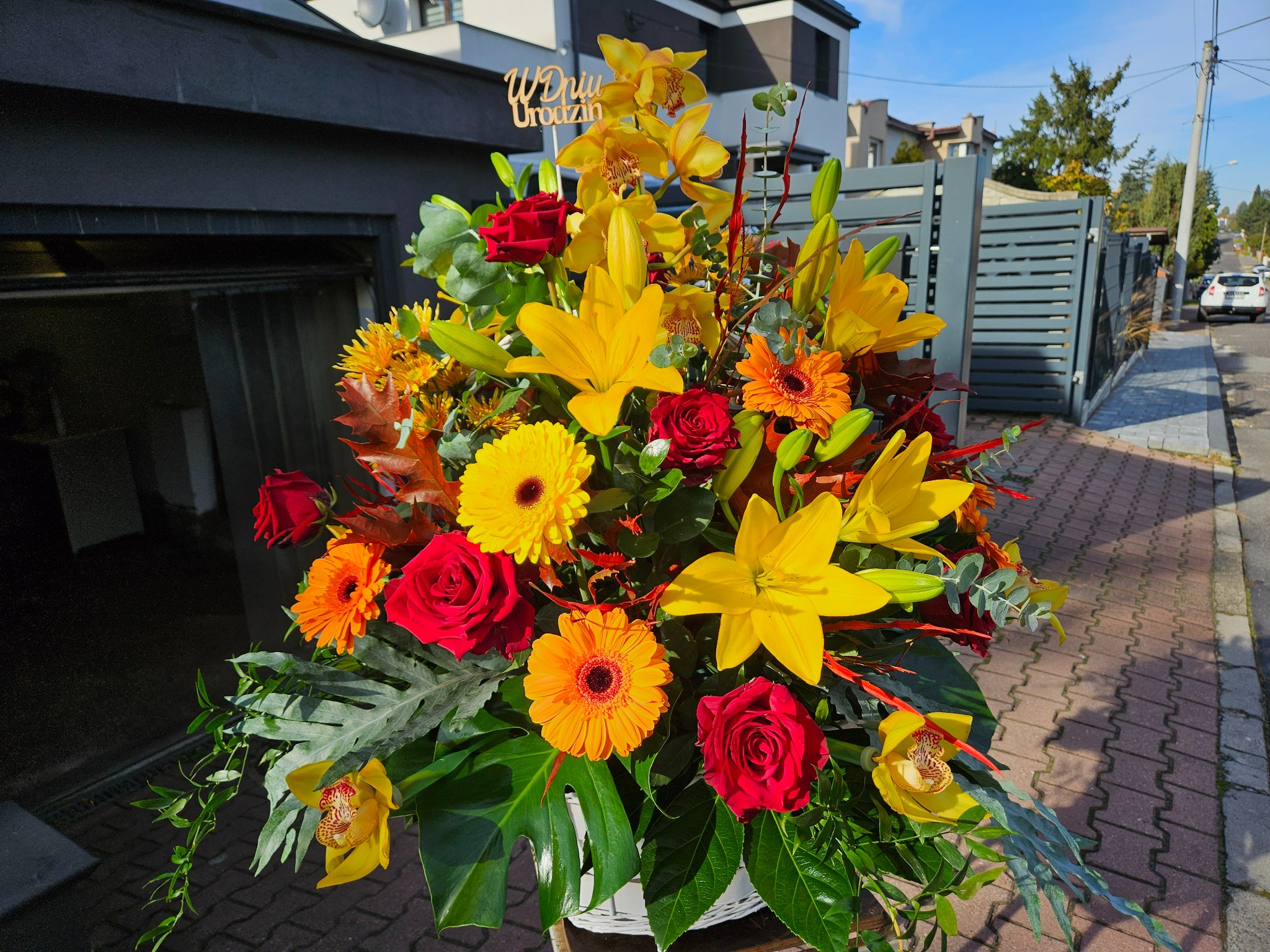 Wielobarwny bukiet z żółtymi liliami, czerwonymi różami i pomarańczowymi gerberami w białym koszyku, z napisem 'W Dniu Urodzin', na tle nowoczesnego domu z szarą bramą i chodnikiem z kostki.