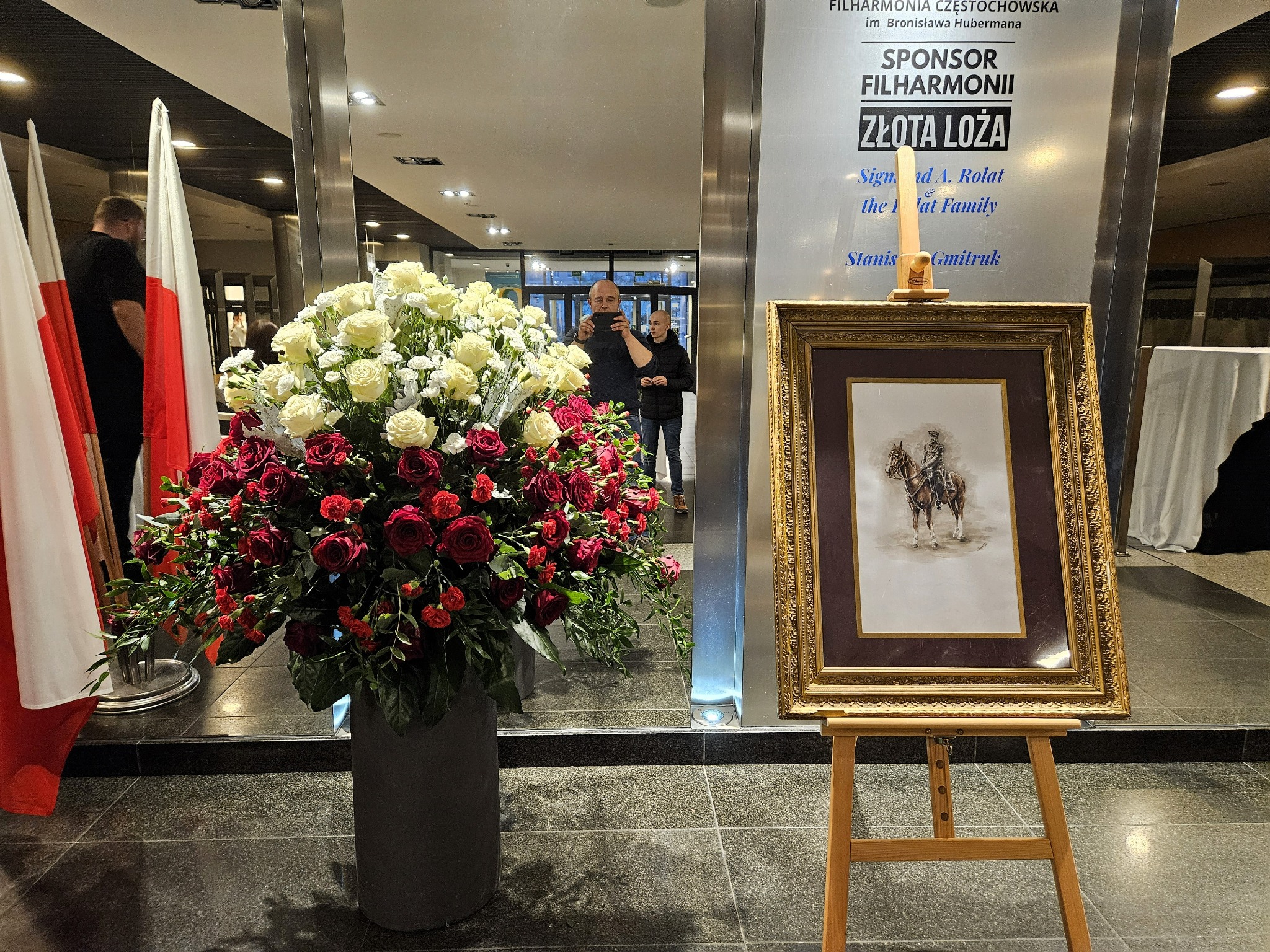 Kompozycja z czerwonych i białych róż w szarej donicy, obok obrazu na sztaludze i flagi Polski w holu Filharmonii Częstochowskiej.