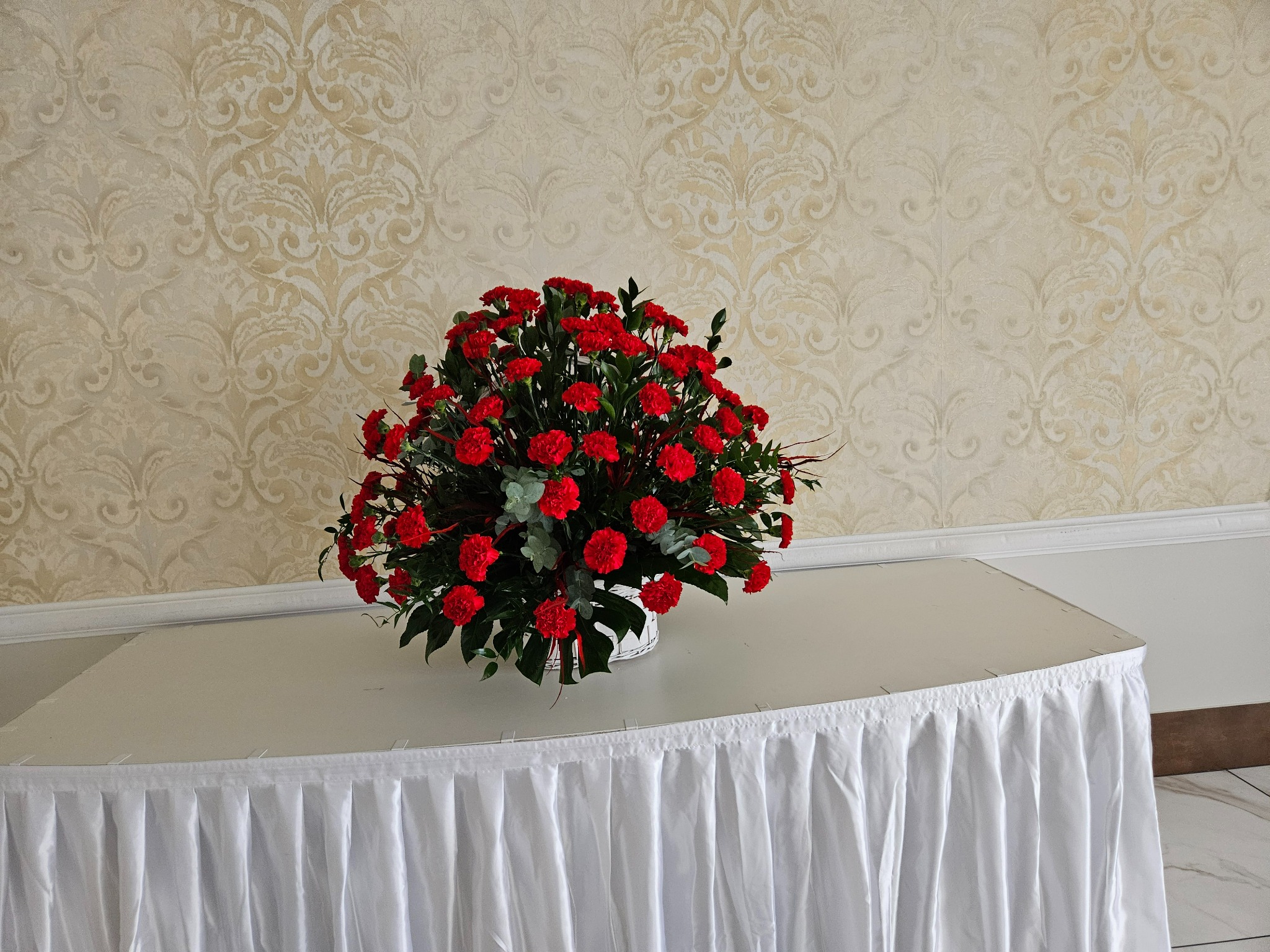 Bogaty bukiet czerwonych goździków z zielonymi liśćmi w białym koszyczku, ustawiony na stole przykrytym białym obrusem z falbanami, na tle ściany z beżową tapetą we wzory.