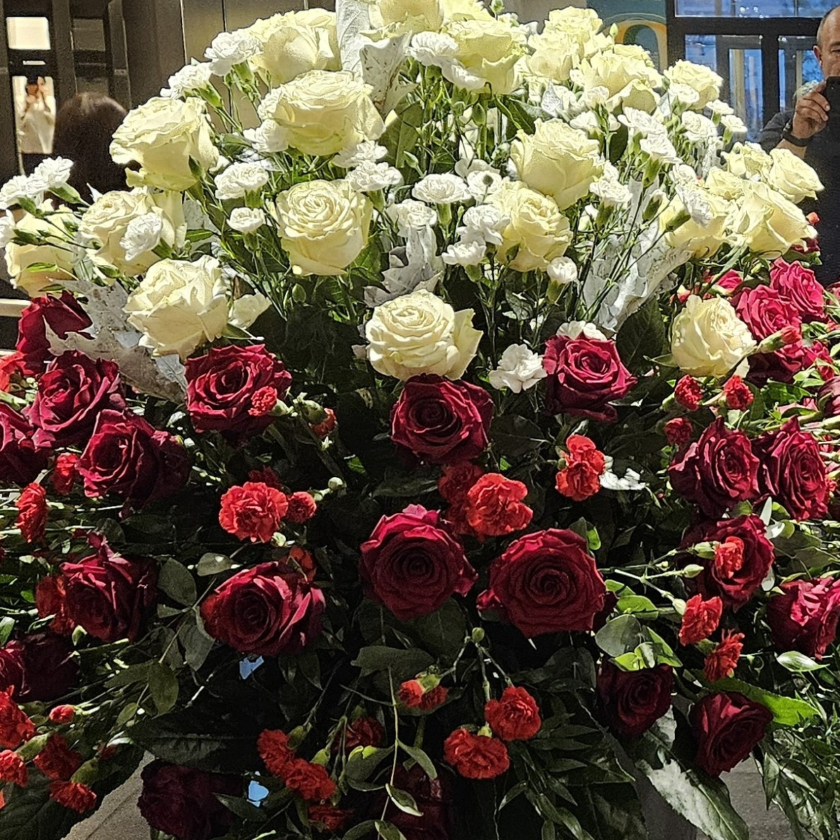 Duży bukiet z białych i czerwonych róż oraz białych goździków, widoczny fragment osoby robiącej zdjęcie telefonem.