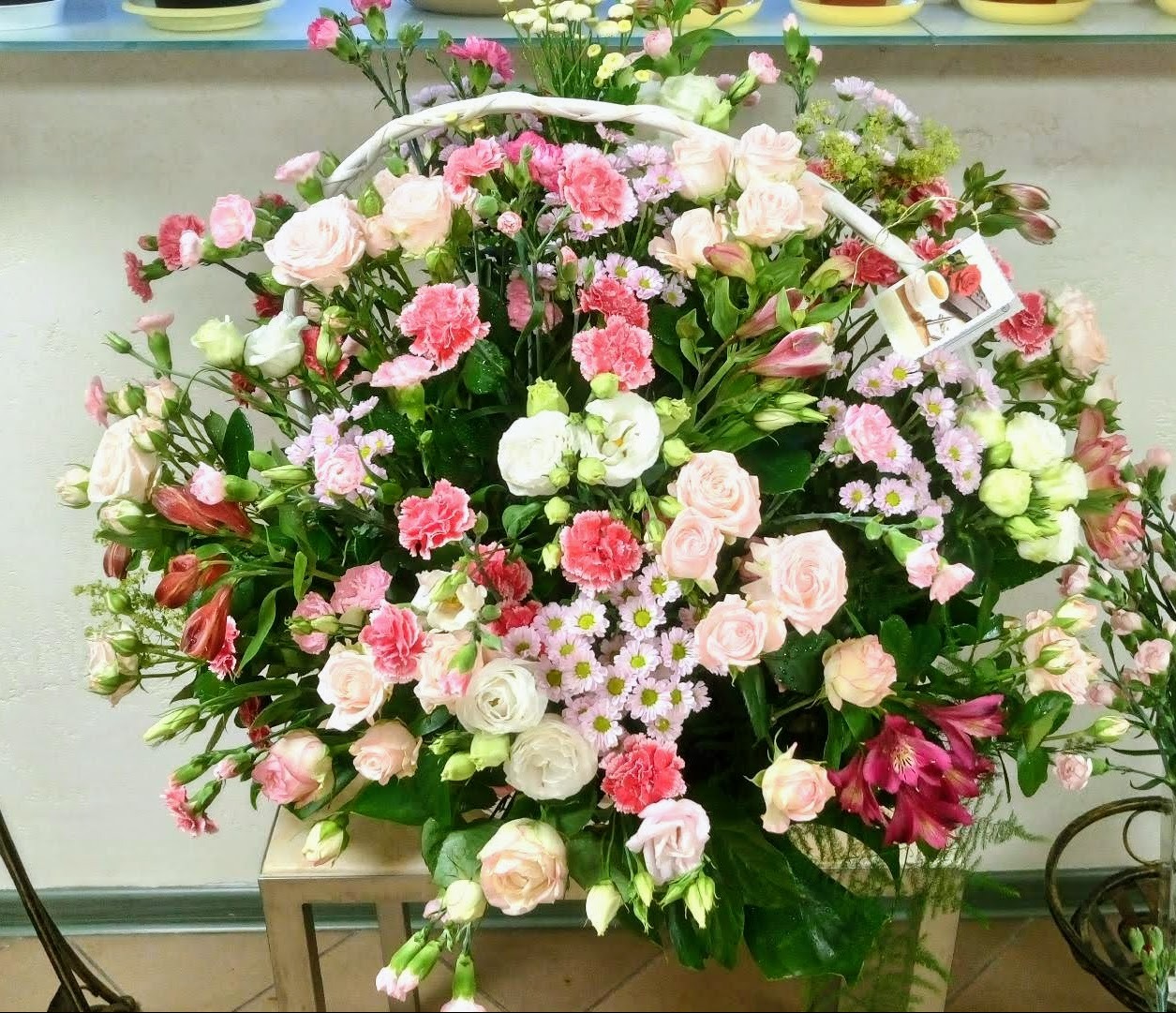 Duży bukiet mieszanych kwiatów w odcieniach różu, bieli i czerwieni, umieszczony w białym koszu, z widoczną kartką z życzeniami.