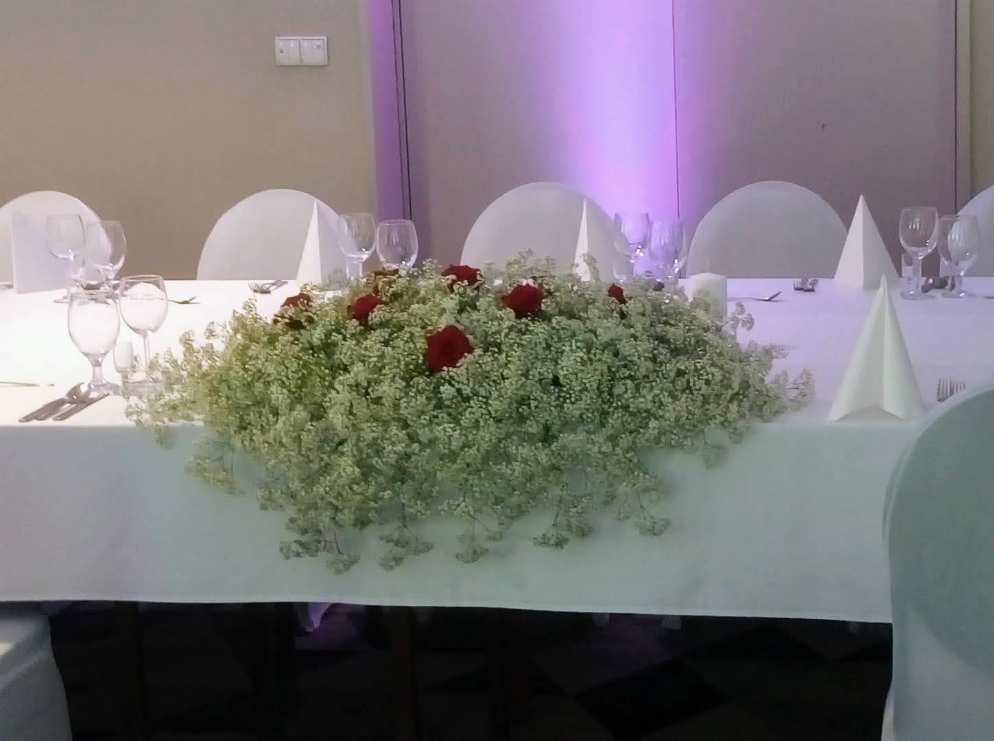 Dekoracja stołu weselnego z białych drobnych kwiatów i czerwonych róż, widoczne nakrycia i złożone serwetki.