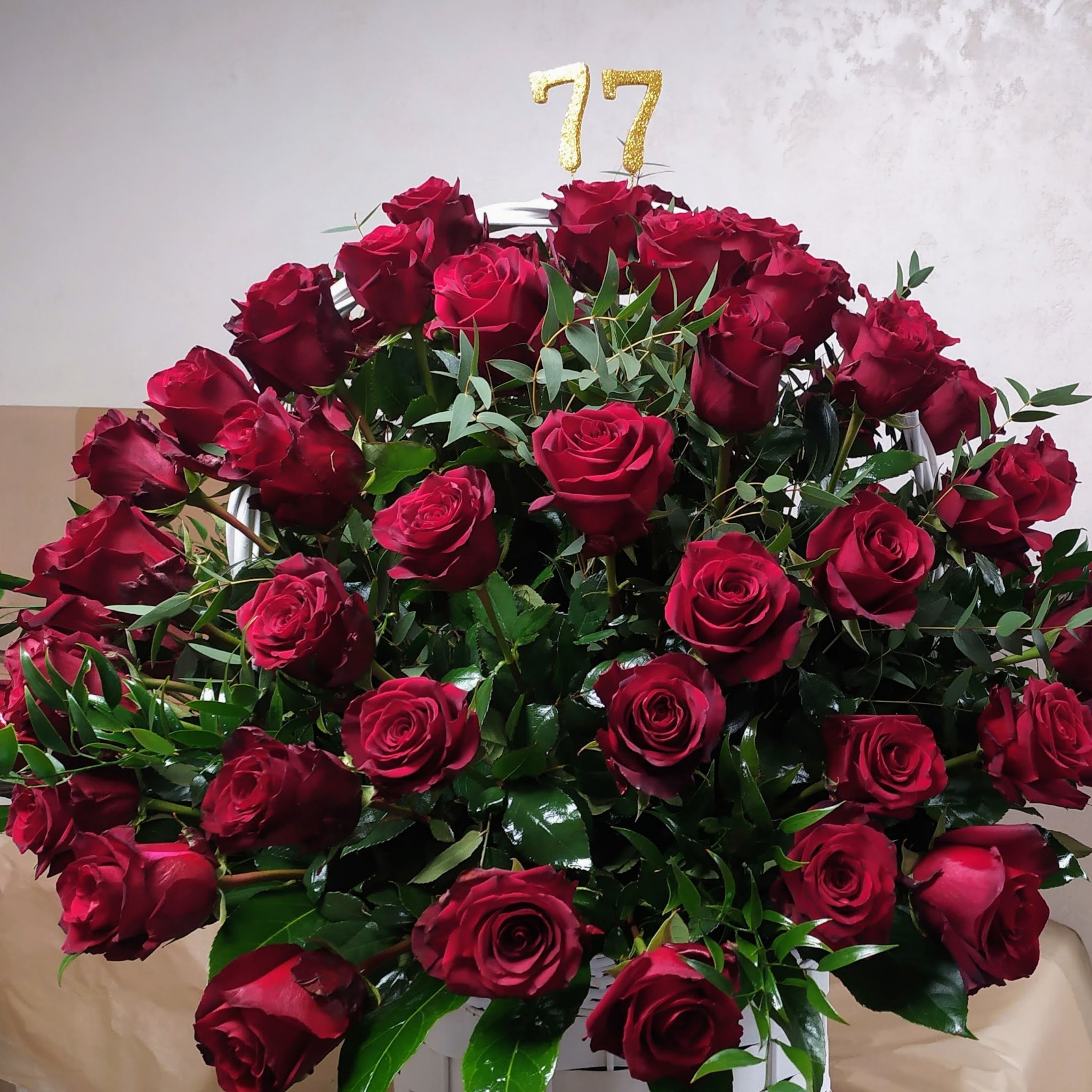 Duży bukiet ciemnoczerwonych róż z zielonymi liśćmi w białym koszu, ozdobiony złotymi cyframi 77.