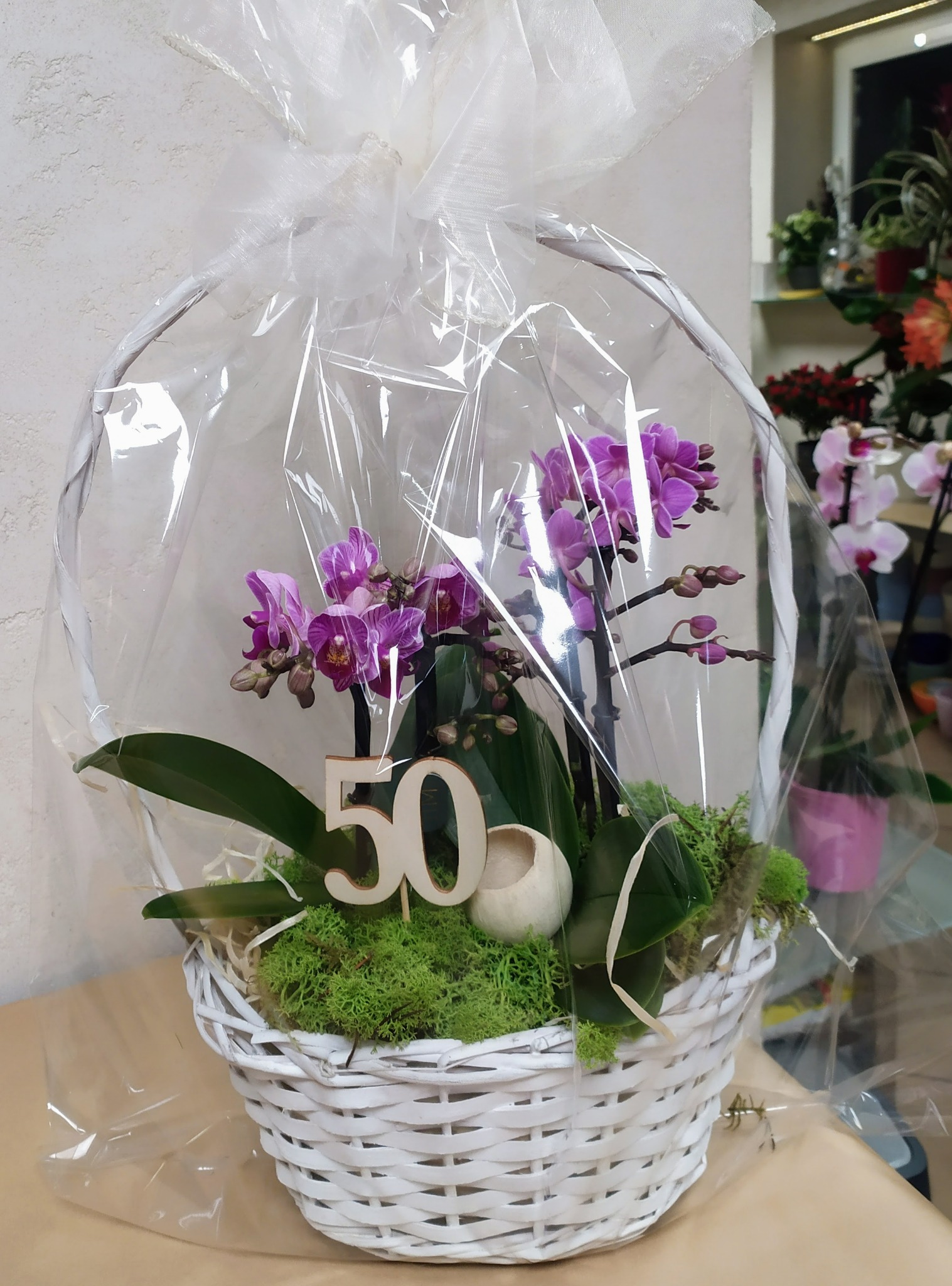 Kosz z fioletowymi storczykami mini, mchem i drewnianą liczbą 50, zapakowany w przezroczysty celofan.