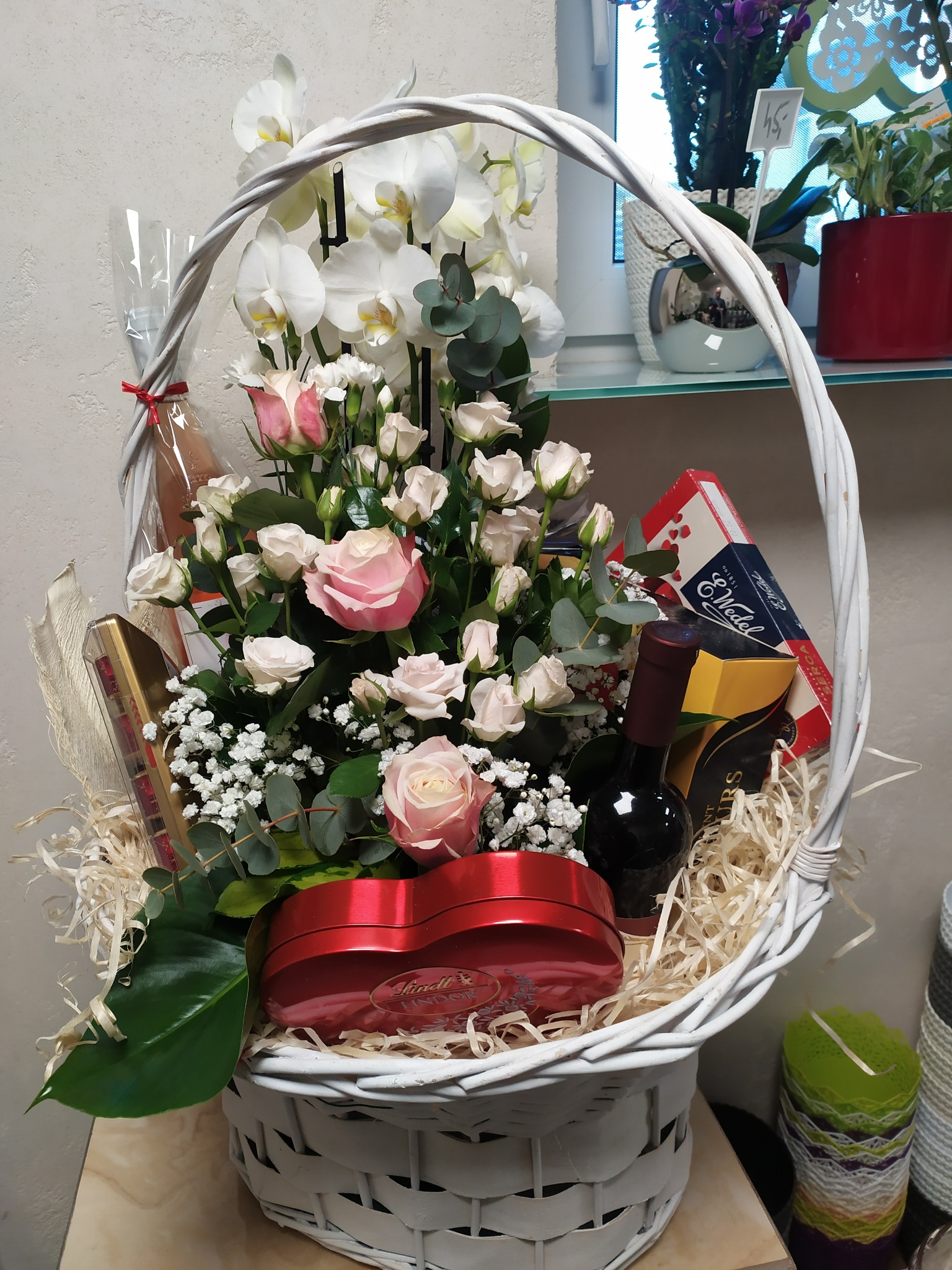 Kosz prezentowy z białymi i różowymi różami, białymi storczykami, butelką wina i słodyczami Lindt oraz Wedel, przygotowany przez kwiaciarnię z Częstochowy.
