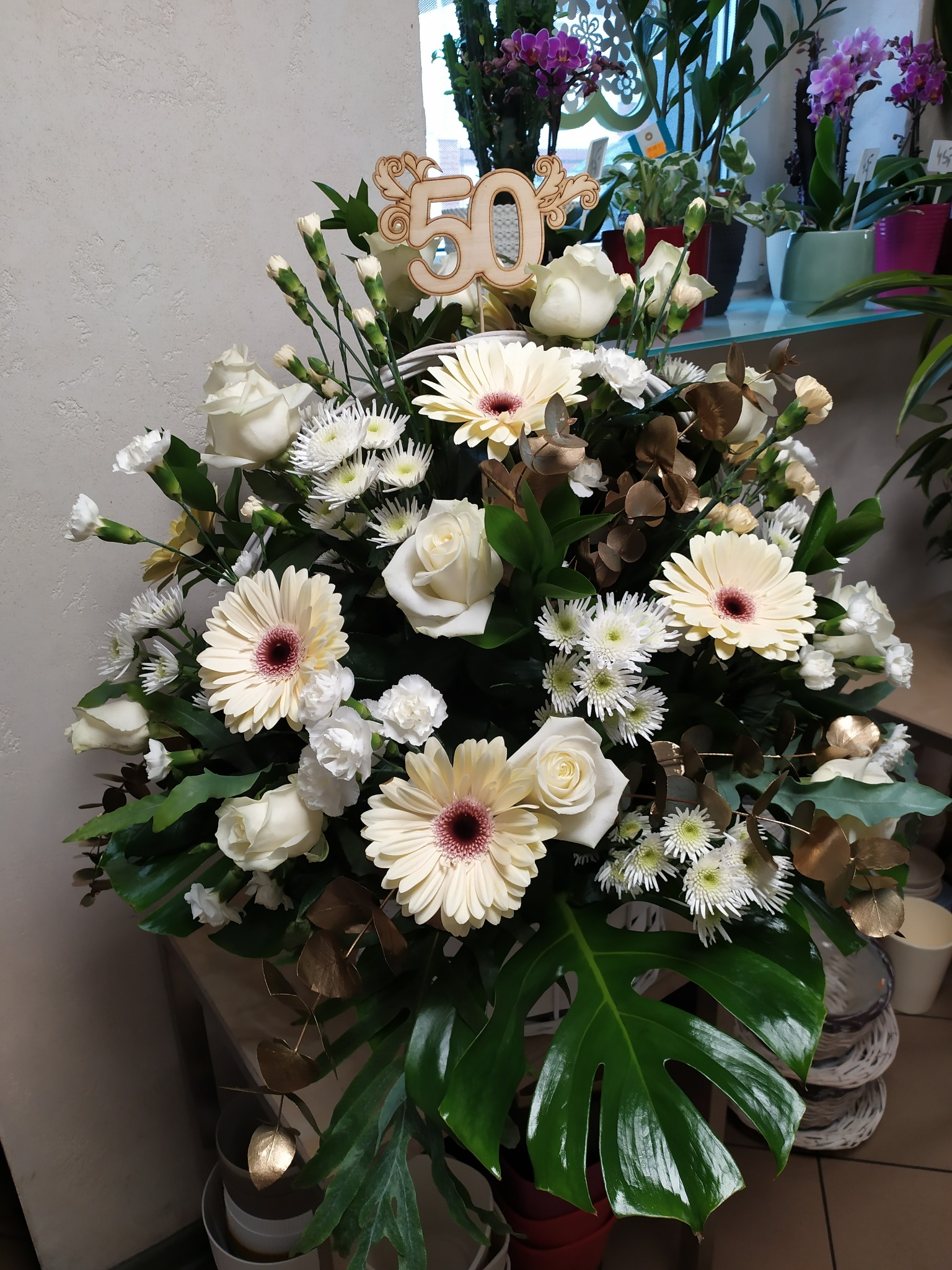 Elegancki bukiet z białych róż, gerber, chryzantem i goździków, ozdobiony drewnianą liczbą 50, na tle roślin doniczkowych na szklanej półce.