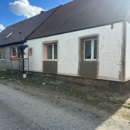 Usługi Budowlane GMB-Bud Rafał Czeszek - Ocieplanie elewacji styropianem: pracownik na rusztowaniu aplikuje zaprawę pod siatkę, widoczne okna z zabezpieczeniem, fragment otynkowanej części przy cokole.