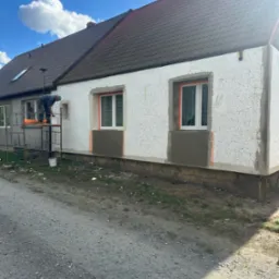 Ocieplanie elewacji styropianem: pracownik na rusztowaniu aplikuje zaprawę pod siatkę, widoczne okna z zabezpieczeniem, fragment otynkowanej części przy cokole.