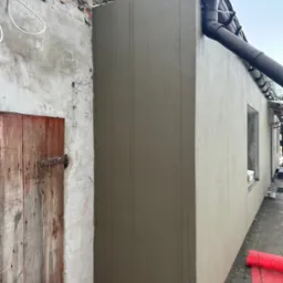 Świeżo otynkowana elewacja budynku z widocznymi śladami po narzędziach, obok stara drewniana brama i fragment ceglanego muru, rynna odprowadzająca wodę deszczową.