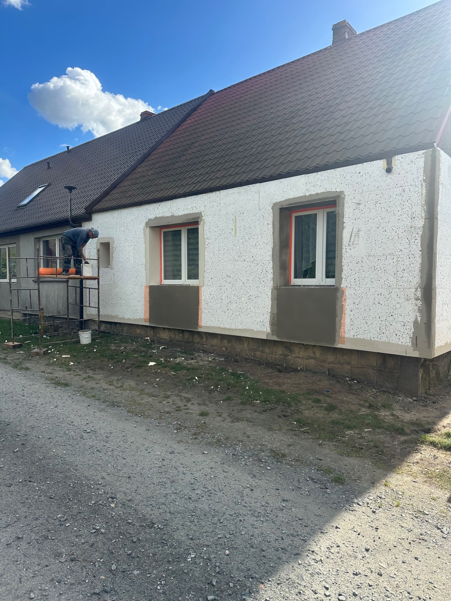 Ocieplanie elewacji styropianem: pracownik na rusztowaniu aplikuje zaprawę pod siatkę, widoczne okna z zabezpieczeniem, fragment otynkowanej części przy cokole.