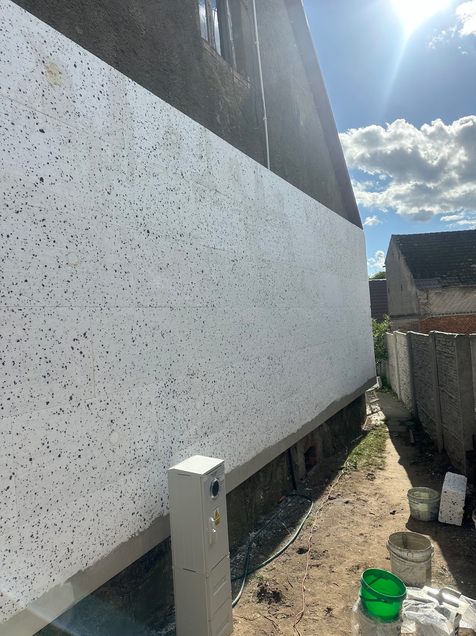 Boczny widok domu z częściowo ocieploną elewacją styropianową, widoczne łączenia płyt, skrzynka elektryczna przy ścianie, wiaderka i narzędzia na ziemi.