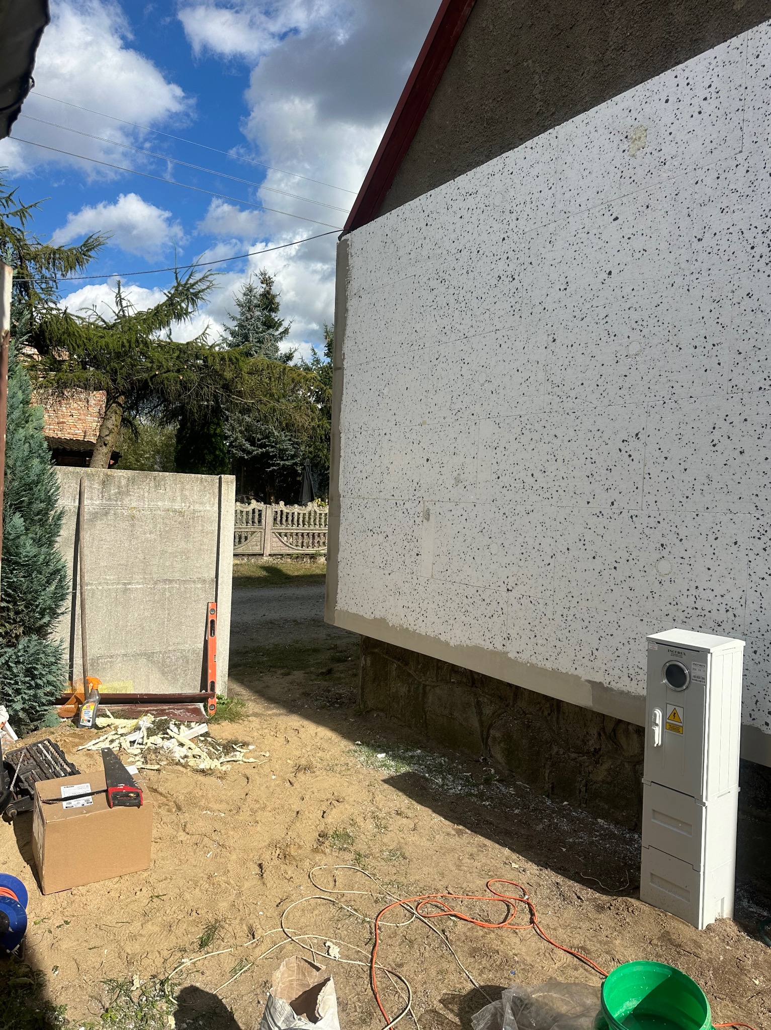 Fragment ocieplonej ściany budynku z widoczną warstwą styropianu, narzędzia i materiały budowlane na ziemi, skrzynka elektryczna przy ścianie, niebo z chmurami w tle.