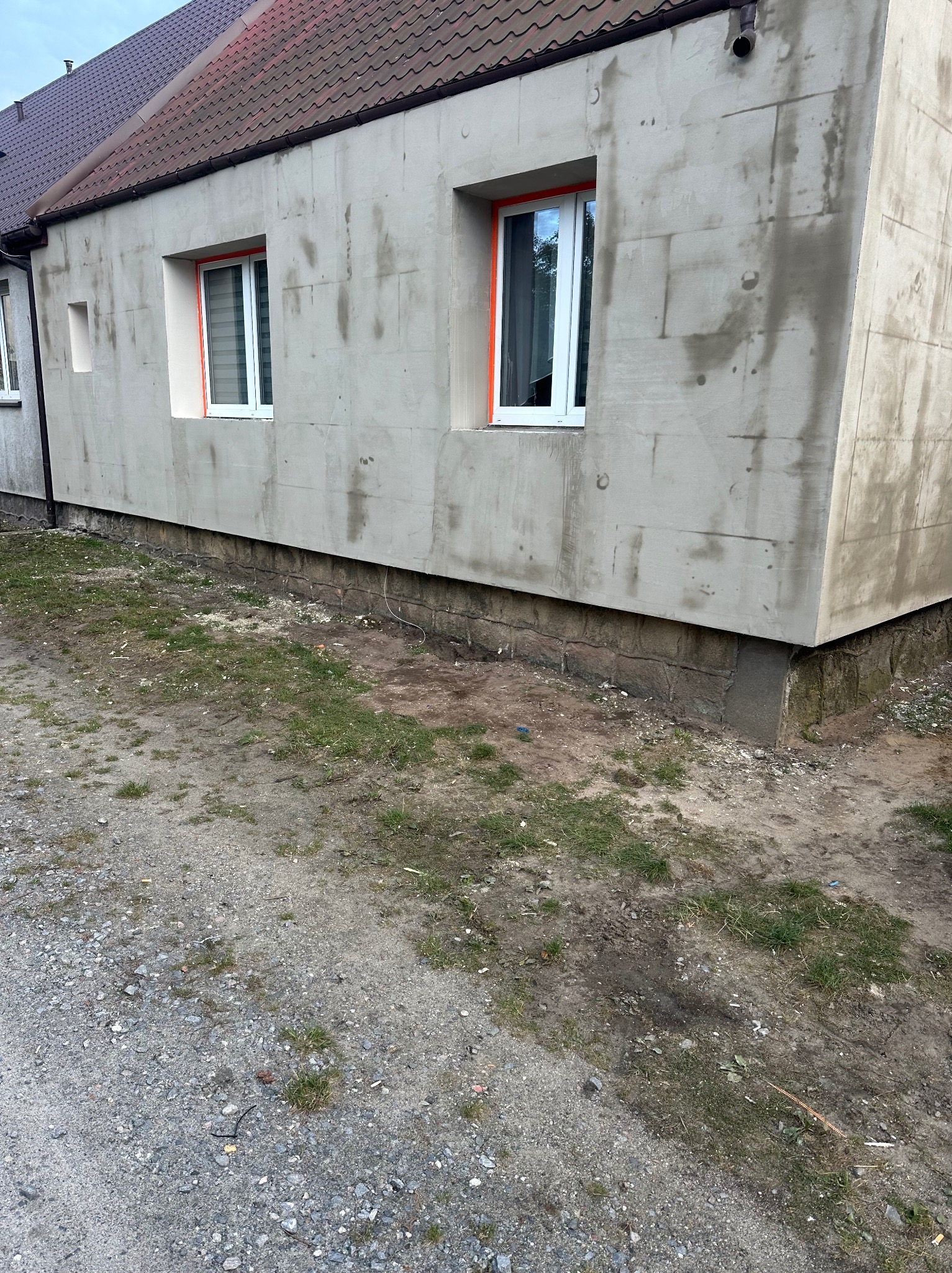 Surowa elewacja domu jednorodzinnego w trakcie prac, widoczne bloki z betonu komórkowego i przygotowane otwory okienne z pomarańczowym zabezpieczeniem, podmurówka z kamienia, teren wokół budynku...