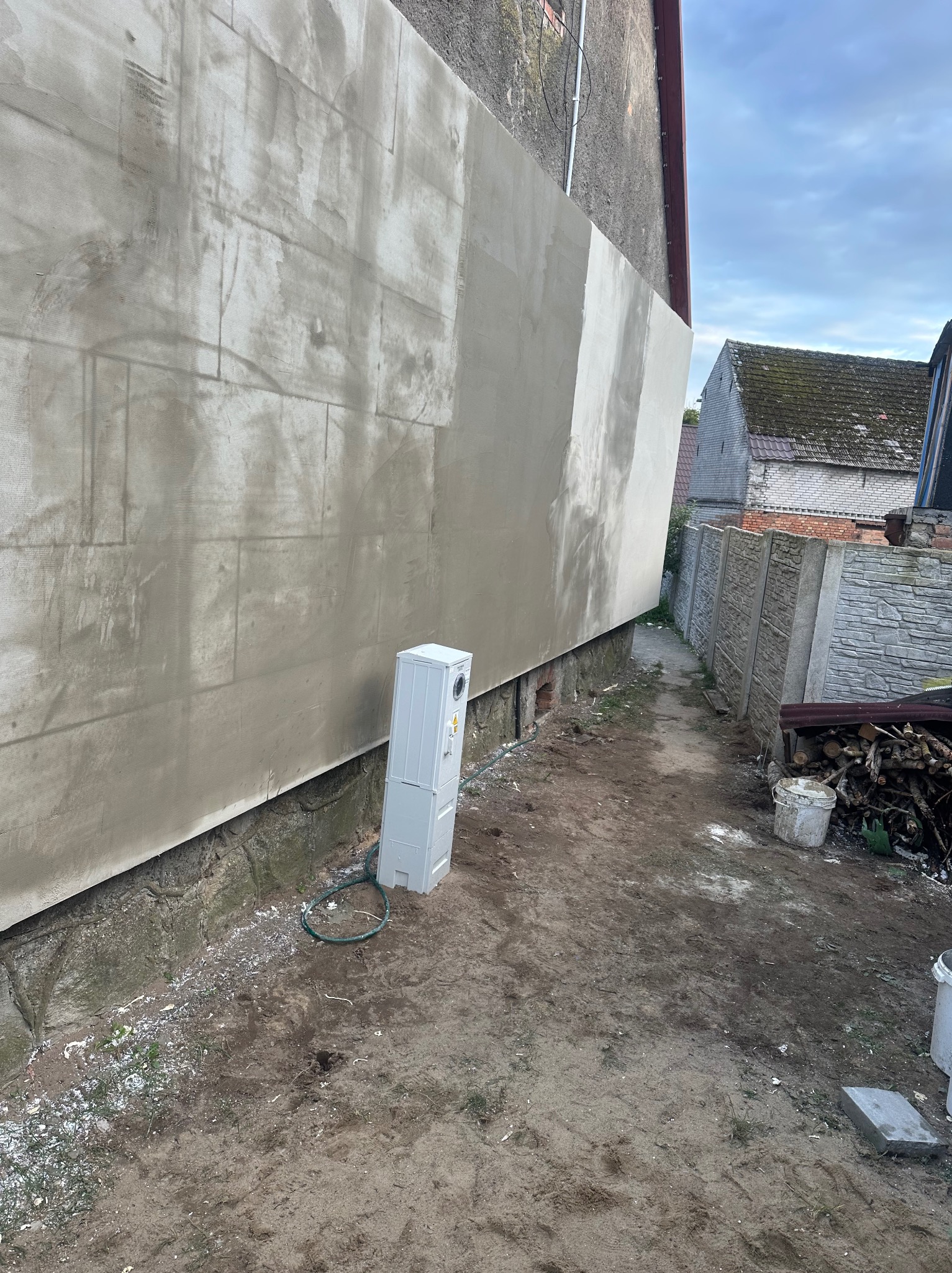 Świeżo otynkowana ściana budynku z widocznymi śladami po łatach, obok skrzynka elektryczna i fragment ogrodu z ziemią i drewnem na opał.
