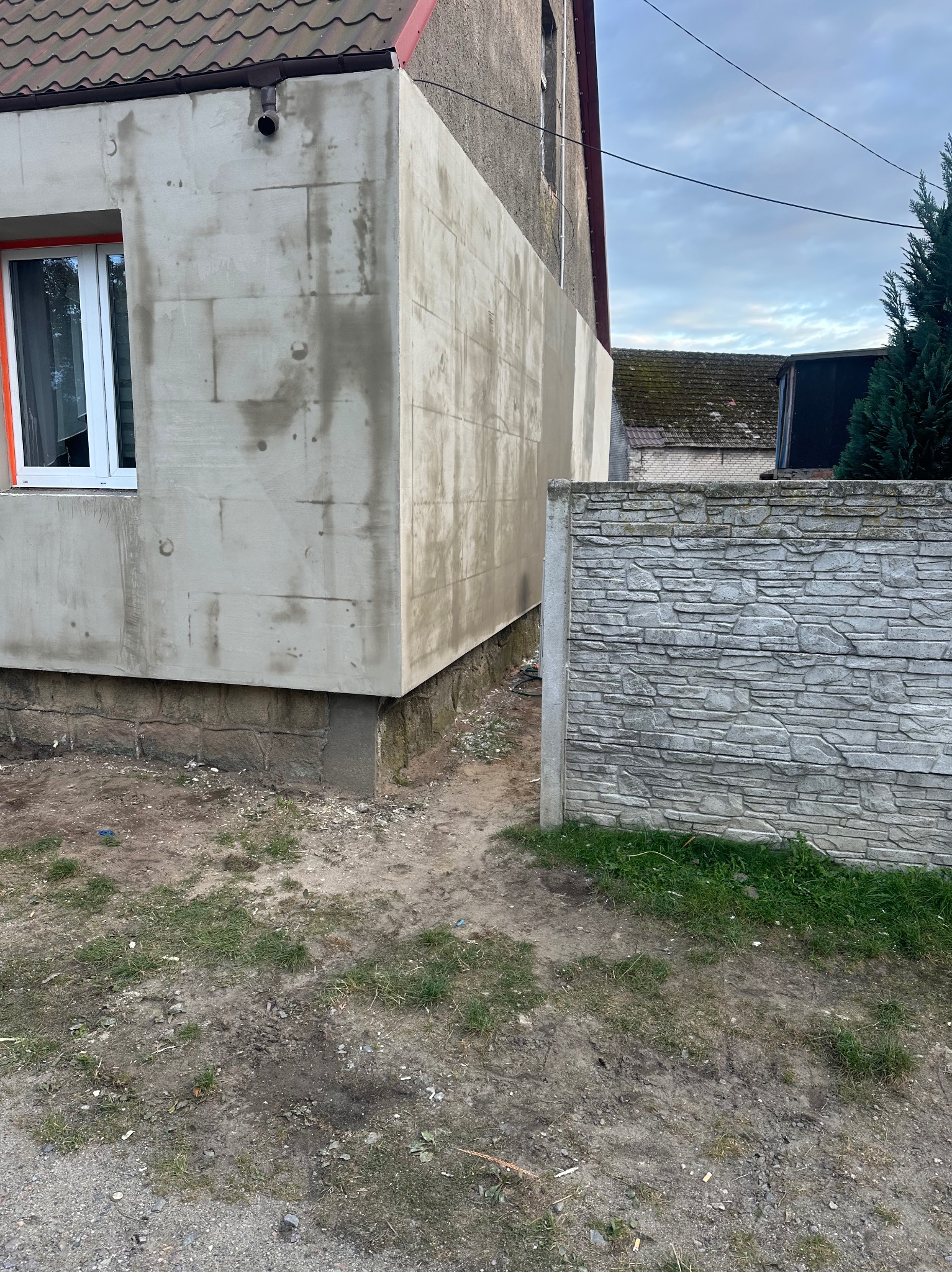 Fragment budynku z ocieploną elewacją, pokrytą szarą warstwą tynku przed nałożeniem warstwy wykończeniowej, widoczne ślady po łatach, okno z pomarańczową ramą, szary betonowy płot w tle.