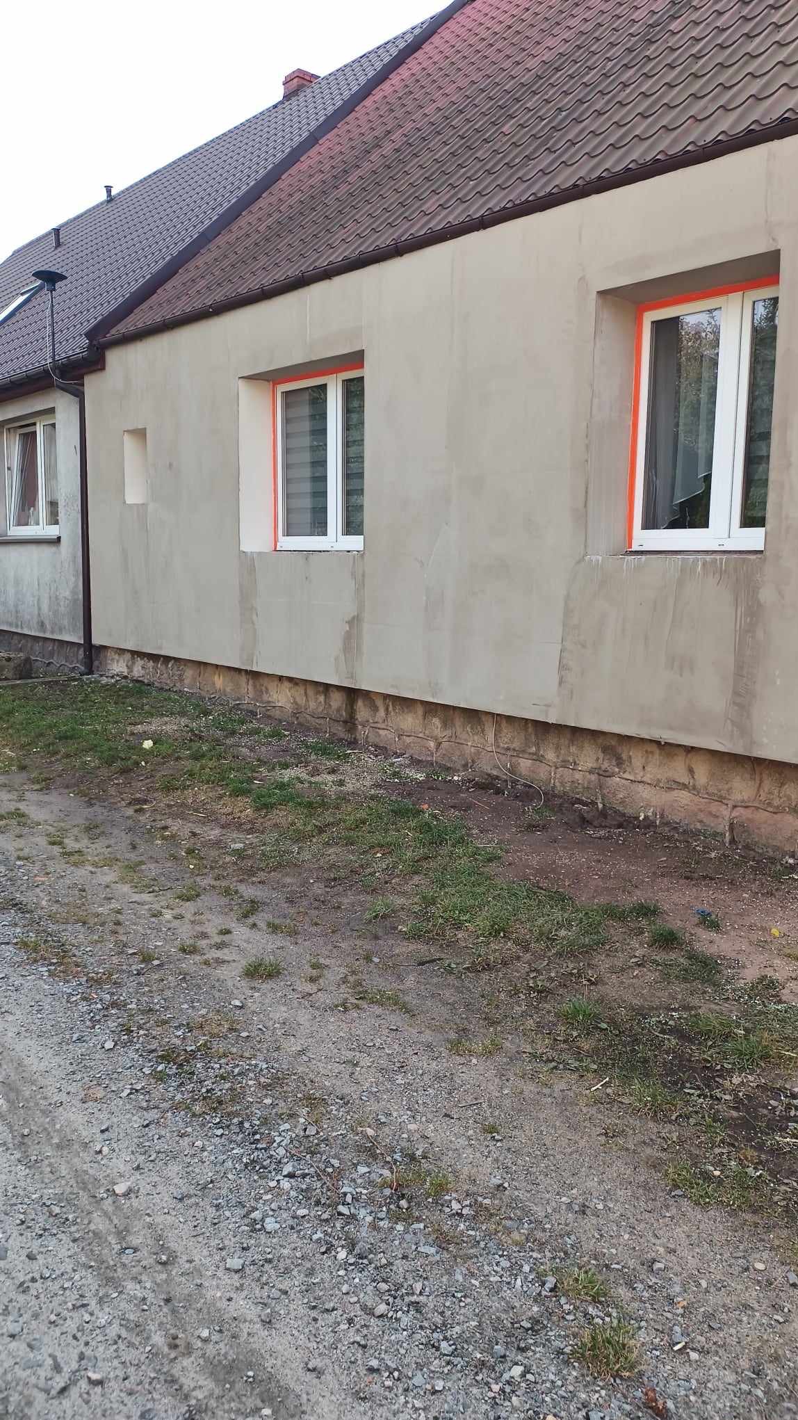 Ściana budynku po nałożeniu szarego tynku, z oknami zabezpieczonymi pomarańczową taśmą malarską, widoczny fragment fundamentu i grunt.