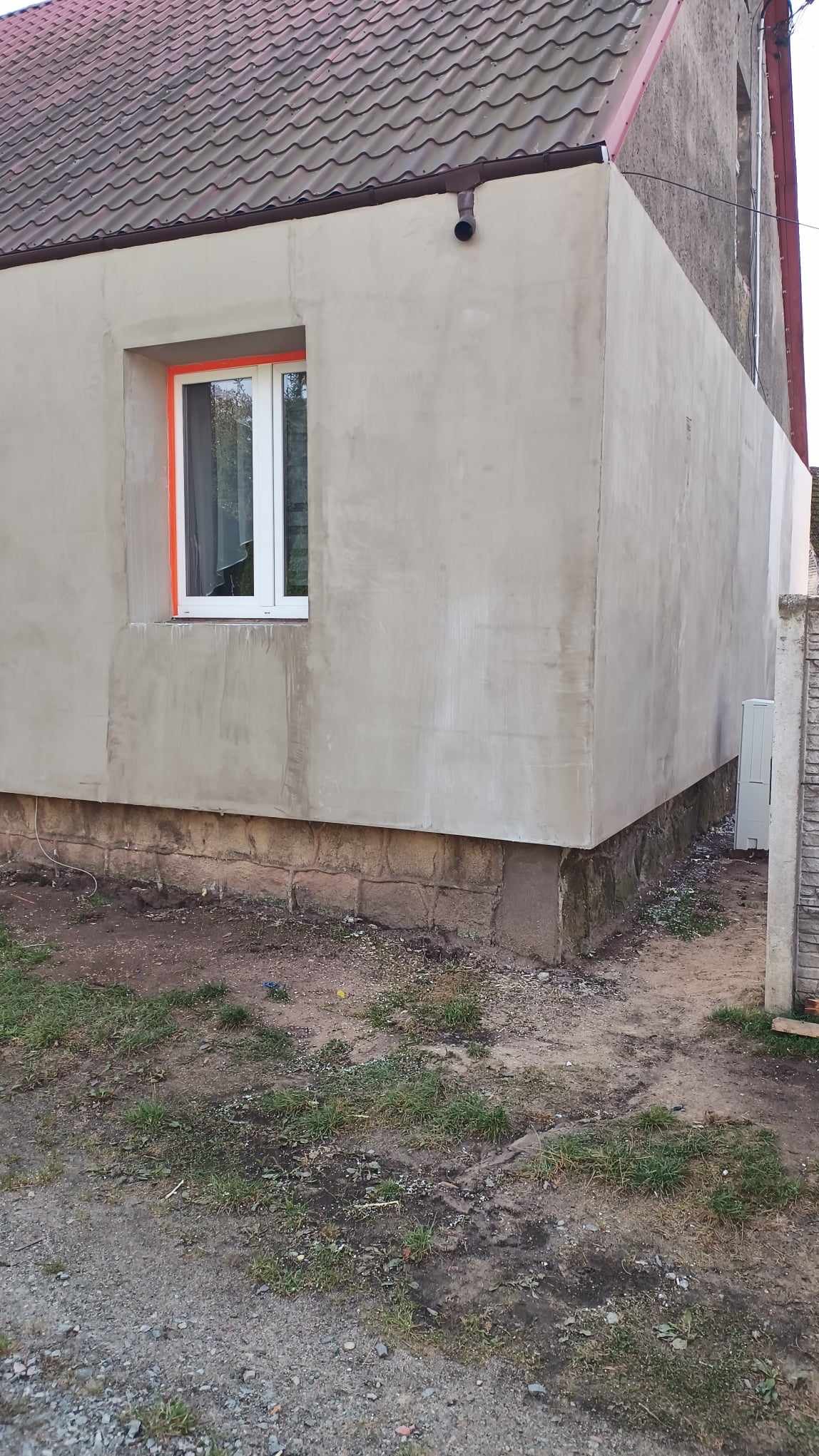 Elewacja budynku po tynkowaniu, z widocznym oknem zabezpieczonym pomarańczową taśmą malarską, oraz fragmentem starej, nieotynkowanej ściany w tle.