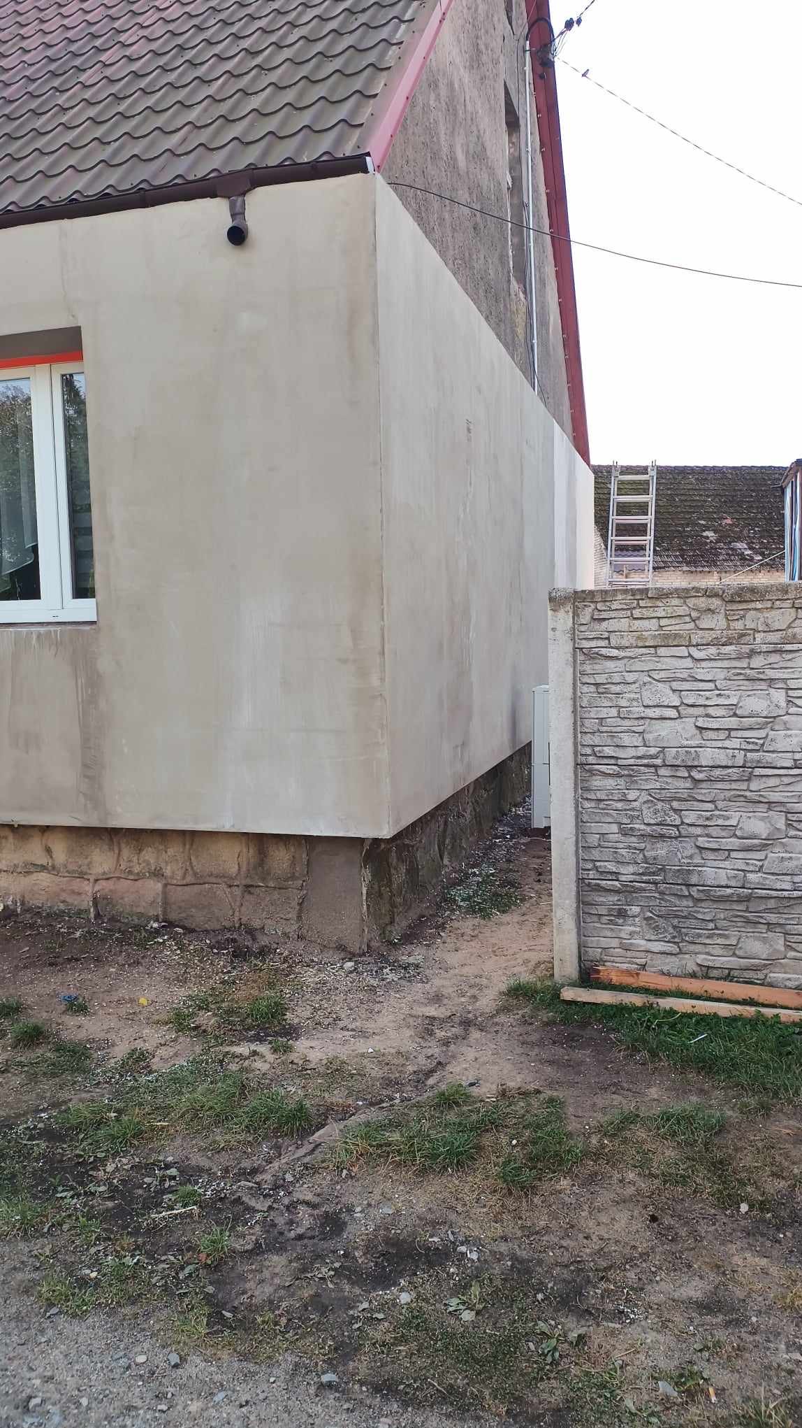 Fragment elewacji domu jednorodzinnego po nałożeniu warstwy tynku, widoczny fragment okna, rynna spustowa i sąsiedni budynek z drabiną opartą o ścianę.