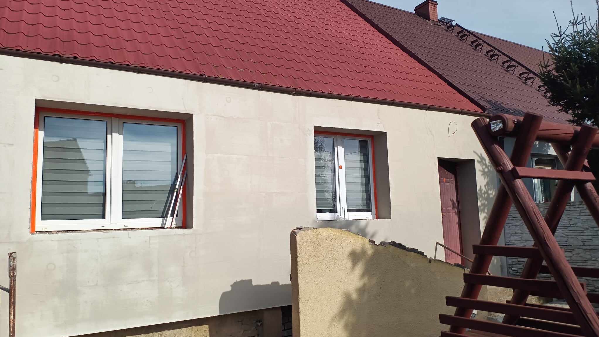 Dom z elewacją w trakcie prac, okna zabezpieczone pomarańczową taśmą, widoczne rusztowanie i fragment drewnianej konstrukcji huśtawki.