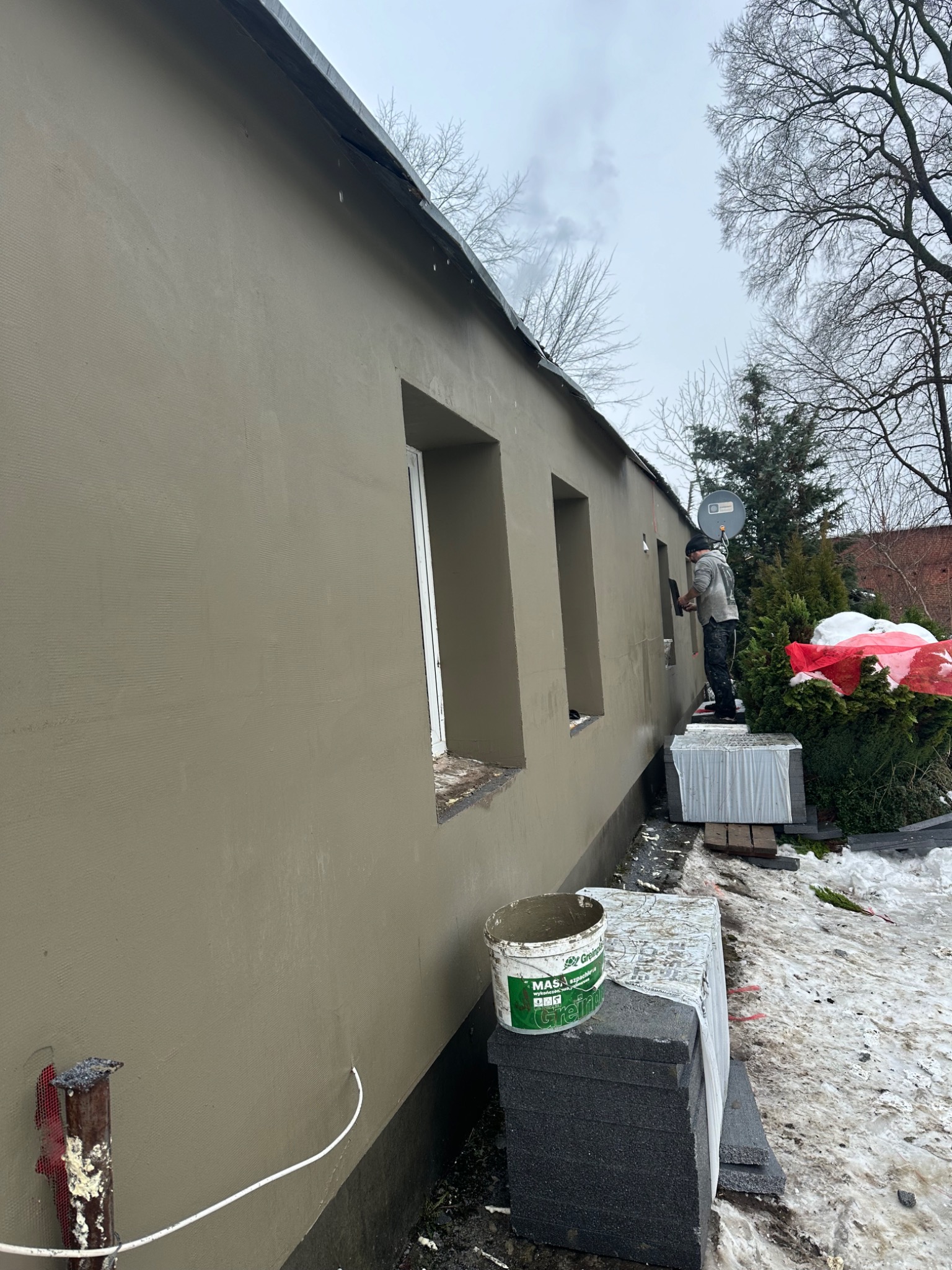 Pracownik w trakcie tynkowania elewacji budynku jednorodzinnego w pochmurny dzień, widoczny kubeł z zaprawą i ocieplenie styropianowe.