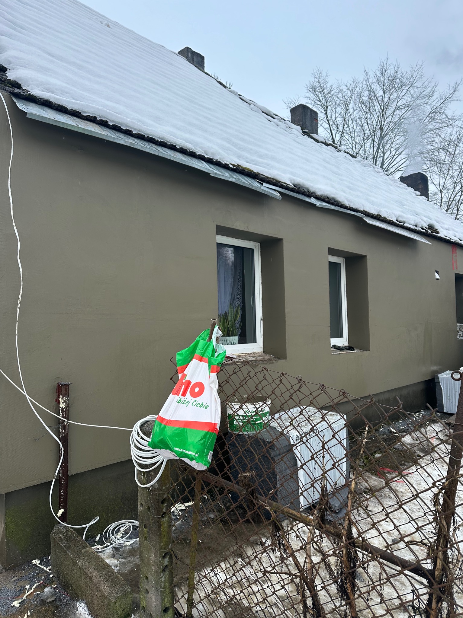 Dom jednorodzinny z elewacją w odcieniach szarości, częściowo pokryty śniegiem, widoczny przez zardzewiałą siatkę ogrodzeniową, z workiem reklamowym i zwiniętym kablem opartym o słupek ogrodzenia.