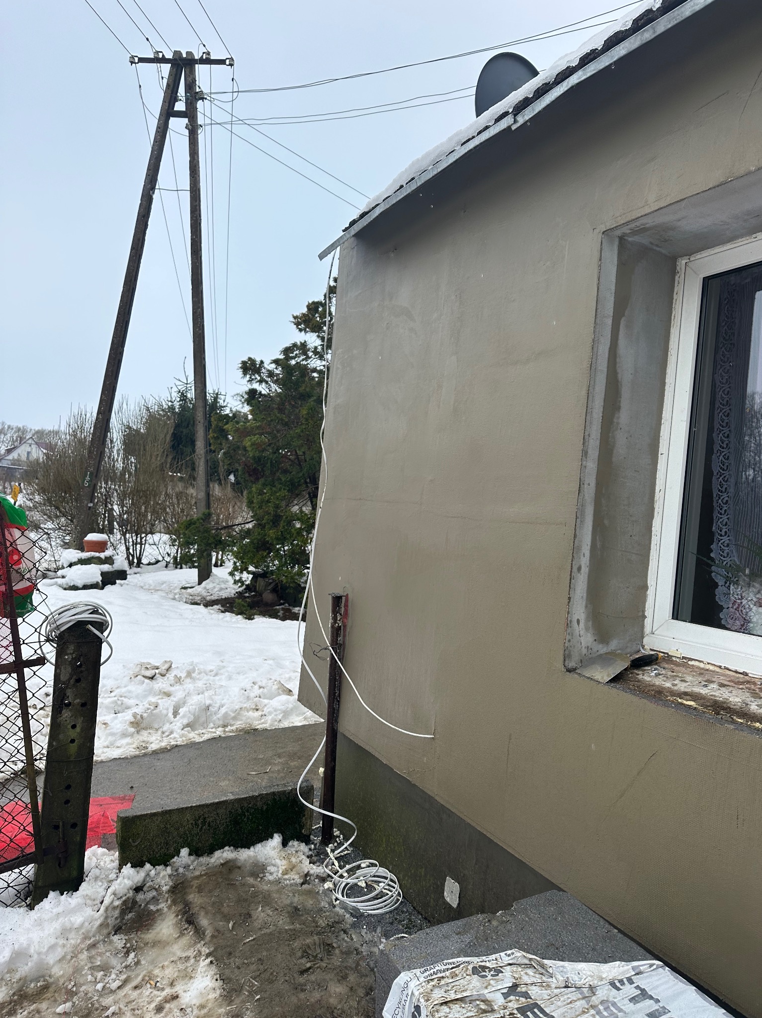 Świeżo otynkowana elewacja domu jednorodzinnego w zimowej scenerii; widoczny fragment okna, kabel zwisający ze ściany oraz worek z materiałem budowlanym