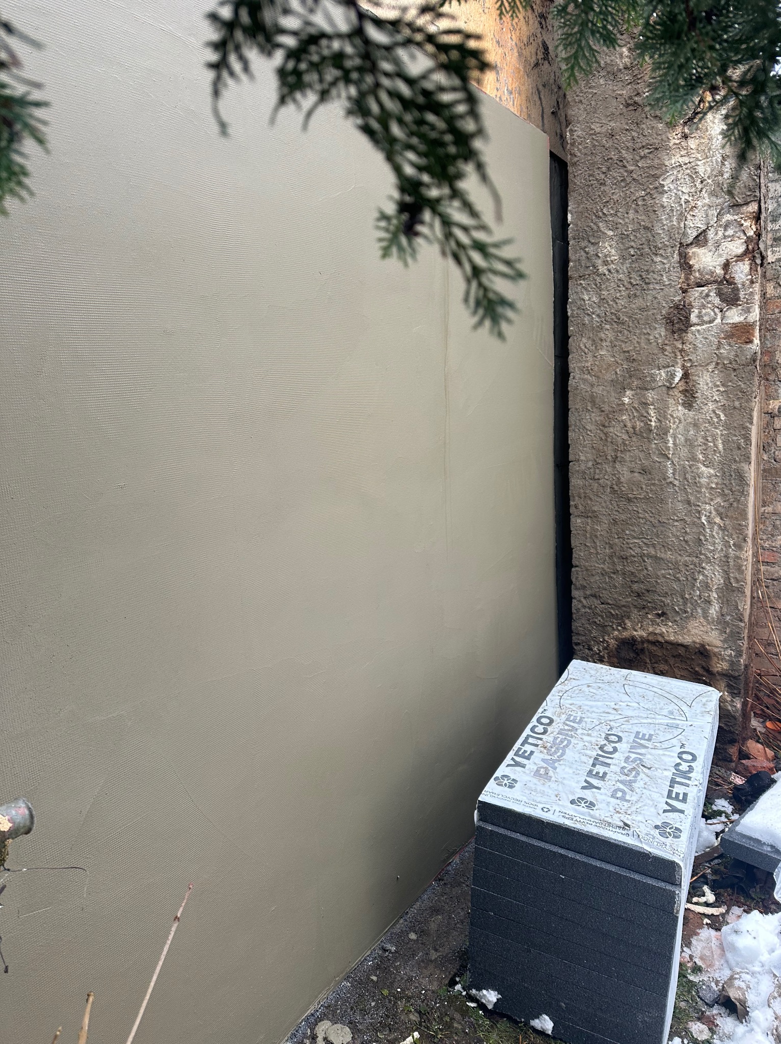 Świeżo otynkowana elewacja budynku przylegająca do starego muru z kamienia, obok leżą płyty styropianowe marki Yetico, na ziemi resztki śniegu.