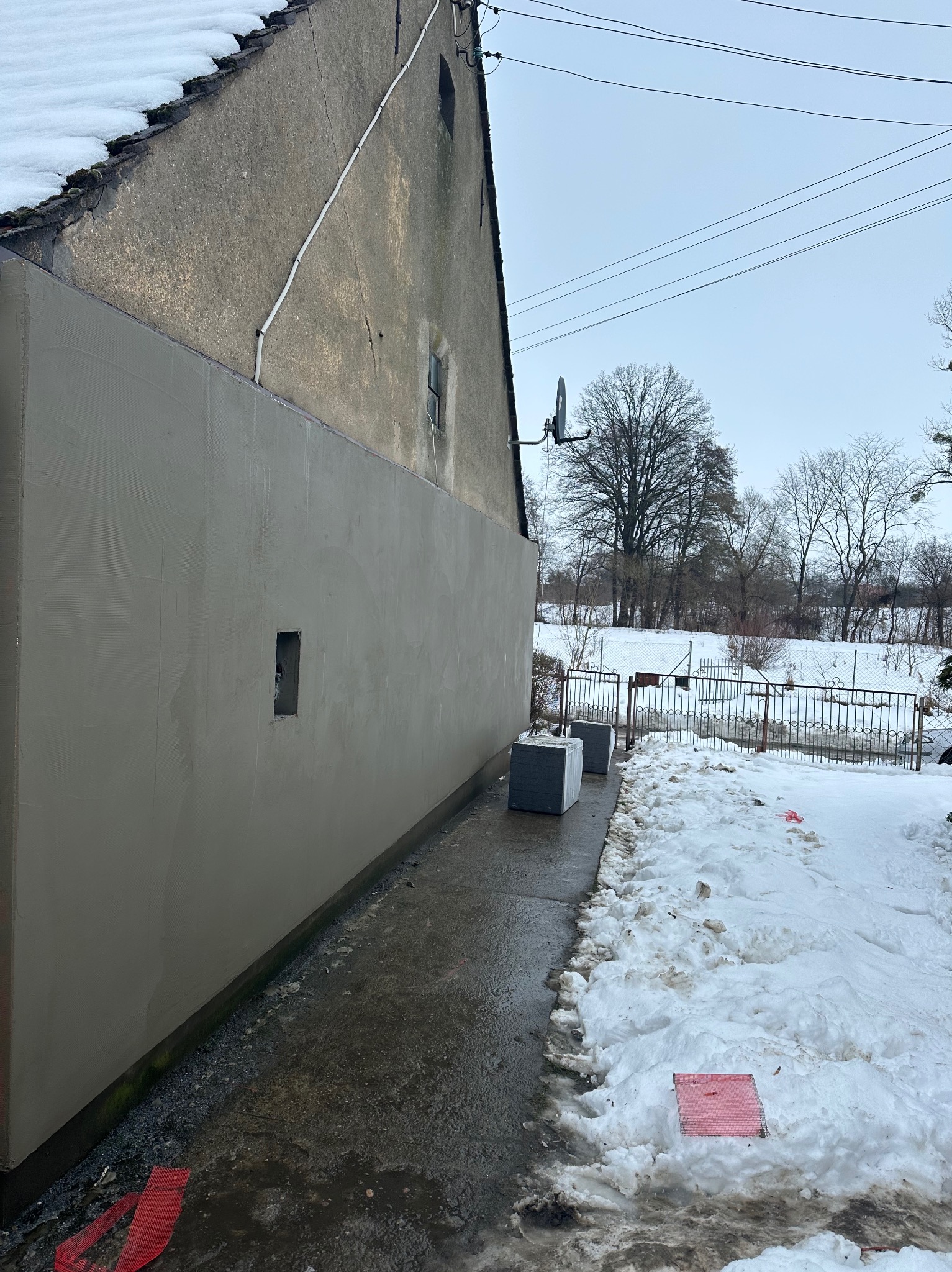Świeżo otynkowana ściana budynku z widocznym okienkiem wentylacyjnym, kostki styropianu obok, zimowy krajobraz w tle.