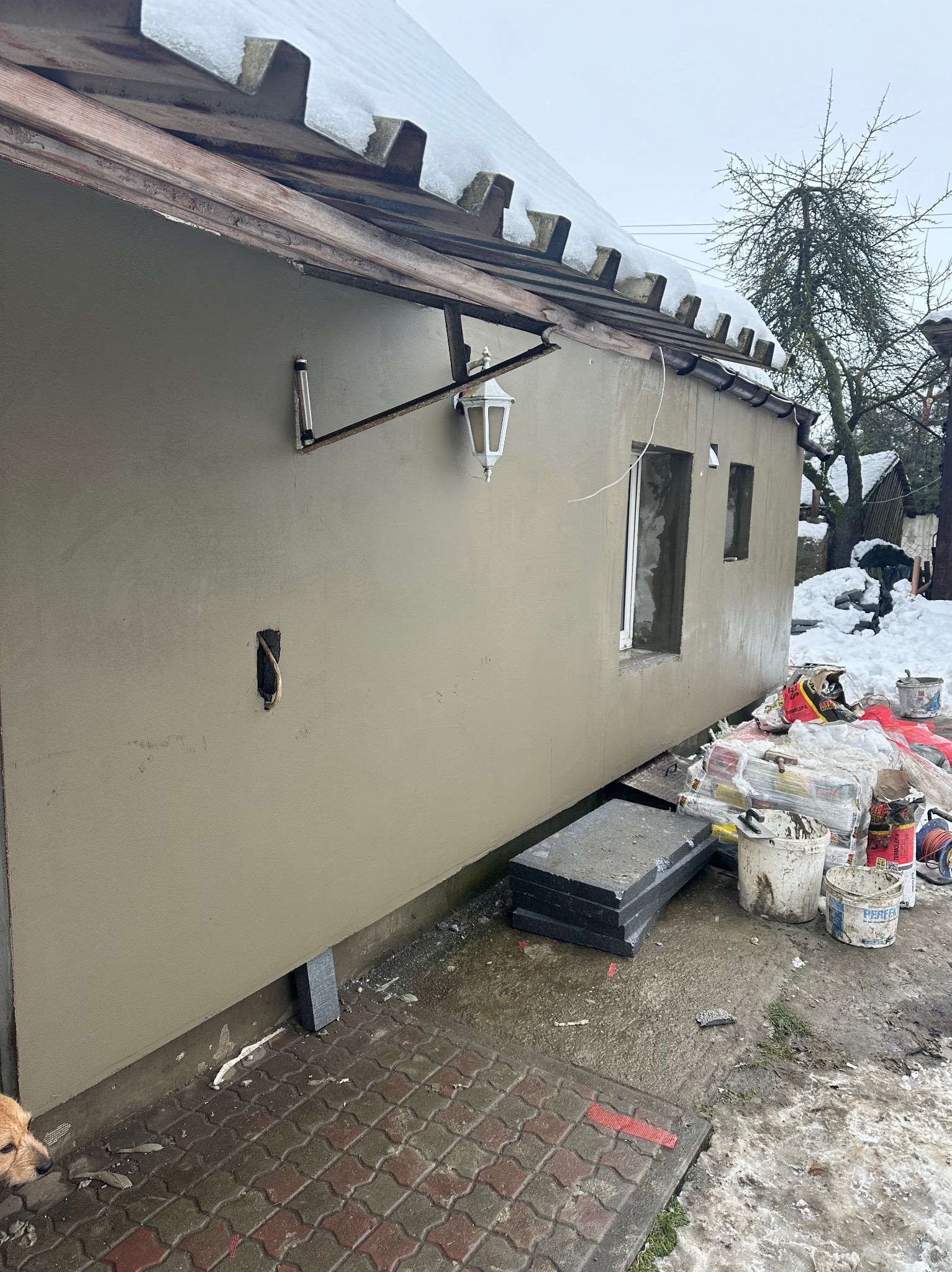 Budynek z nową, gładką, beżową elewacją w trakcie prac wykończeniowych, widoczne puszki z materiałami, ocieplenie i wystający kabel elektryczny, na dachu śnieg.