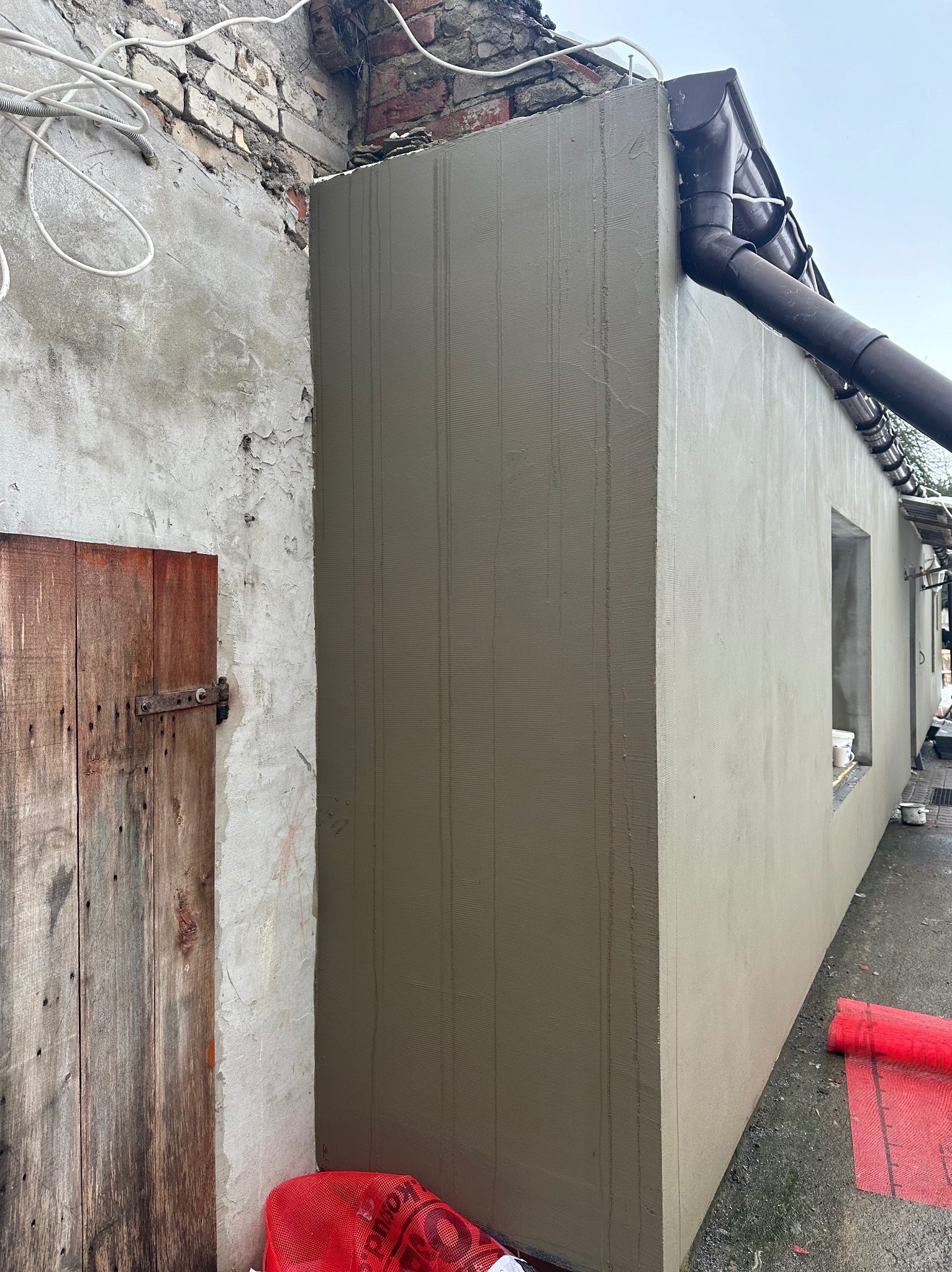 Świeżo otynkowana elewacja budynku z widocznymi śladami po narzędziach, obok stara drewniana brama i fragment ceglanego muru, rynna odprowadzająca wodę deszczową.