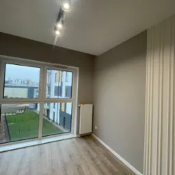 Pusty pokój z panelami ściennymi, oknem z widokiem na osiedle i lampą sufitową z pięcioma reflektorami.