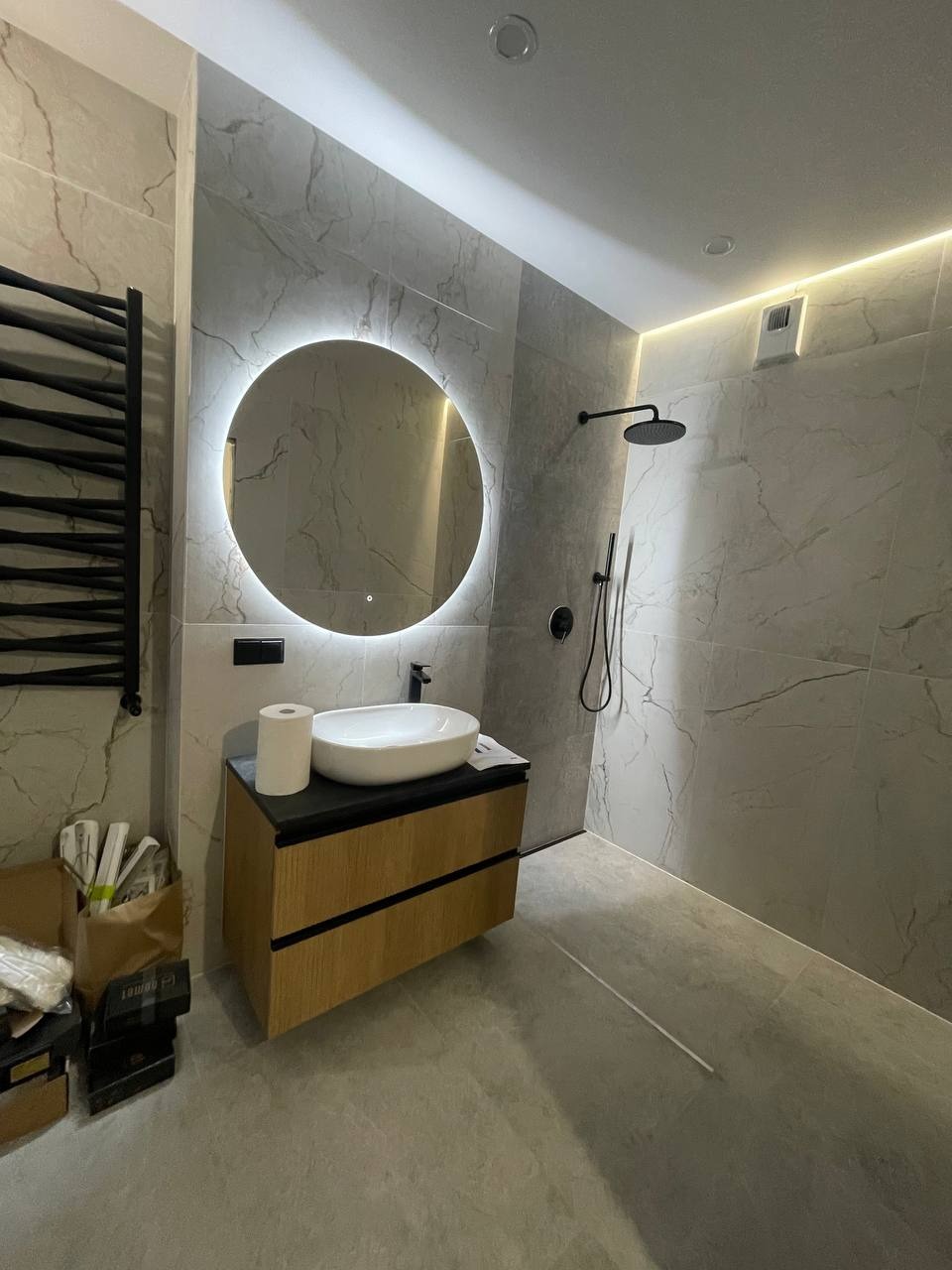 Nowoczesna łazienka z okrągłym lustrem podświetlanym LED, białą umywalką nablatową na drewnianej szafce z czarnym blatem, szarymi płytkami imitującymi marmur i czarną armaturą.