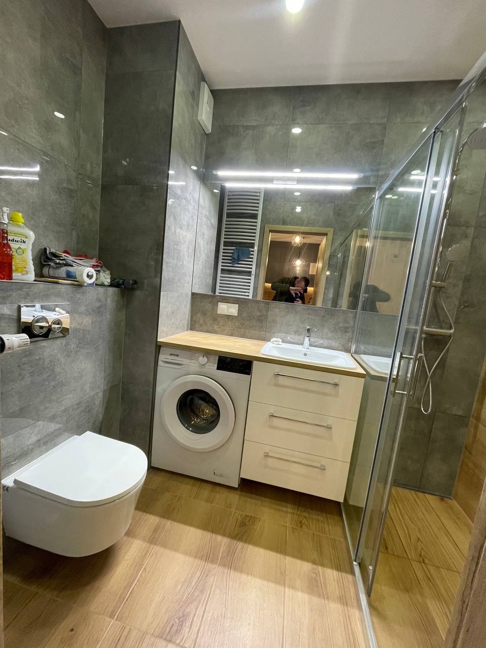 Nowoczesna łazienka z szarymi płytkami imitującymi kamień, drewnopodobną podłogą, białą toaletą, pralką pod blatem i kabiną prysznicową ze szklanymi drzwiami.