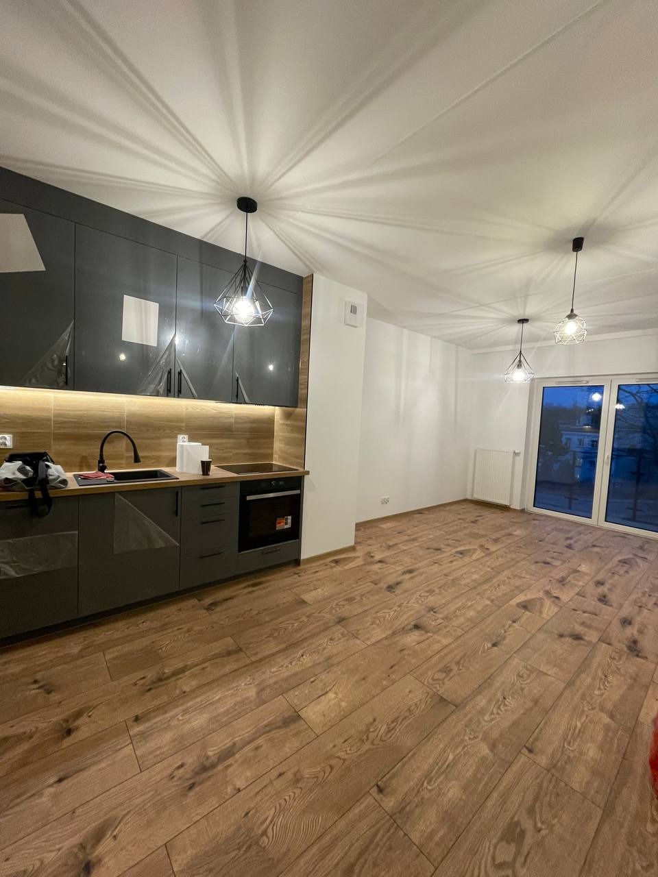 Nowoczesna kuchnia z szarymi szafkami, drewnianym blatem i podłogą z paneli, oświetlona trzema geometrycznymi lampami wiszącymi, z widokiem na zewnątrz przez przesuwne drzwi balkonowe.