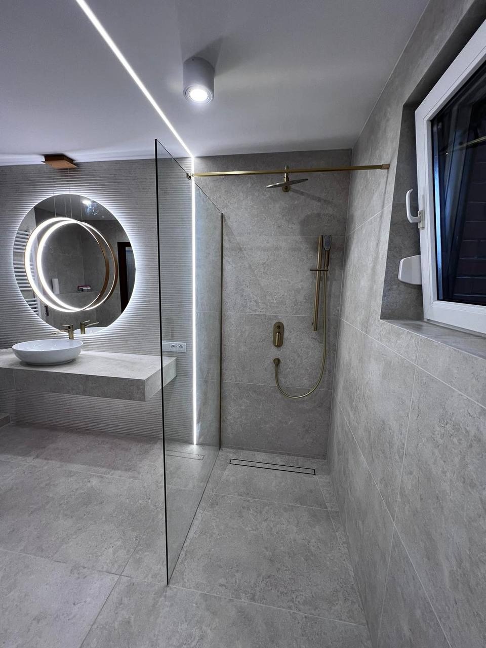 Nowoczesna łazienka w odcieniach szarości z elementami złota: okrągłe lustro z podświetleniem LED, szklana kabina prysznicowa ze złotą armaturą i odpływem liniowym, umywalka nablatowa na szarej...