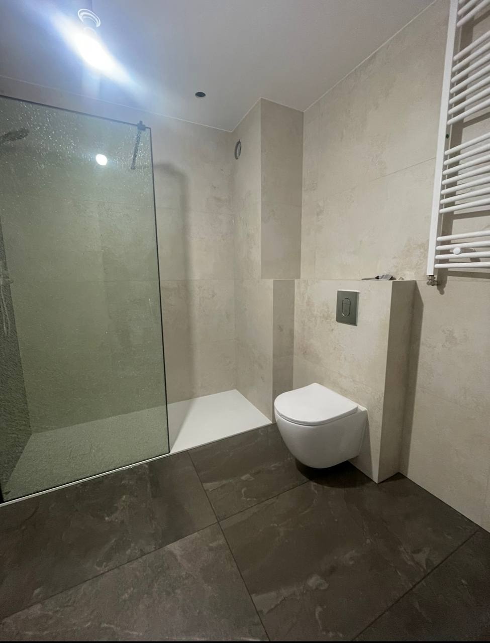 Nowoczesna łazienka z kabiną prysznicową typu walk-in, wiszącą toaletą i grzejnikiem drabinkowym na beżowych płytkach ściennych i ciemnych płytkach podłogowych.